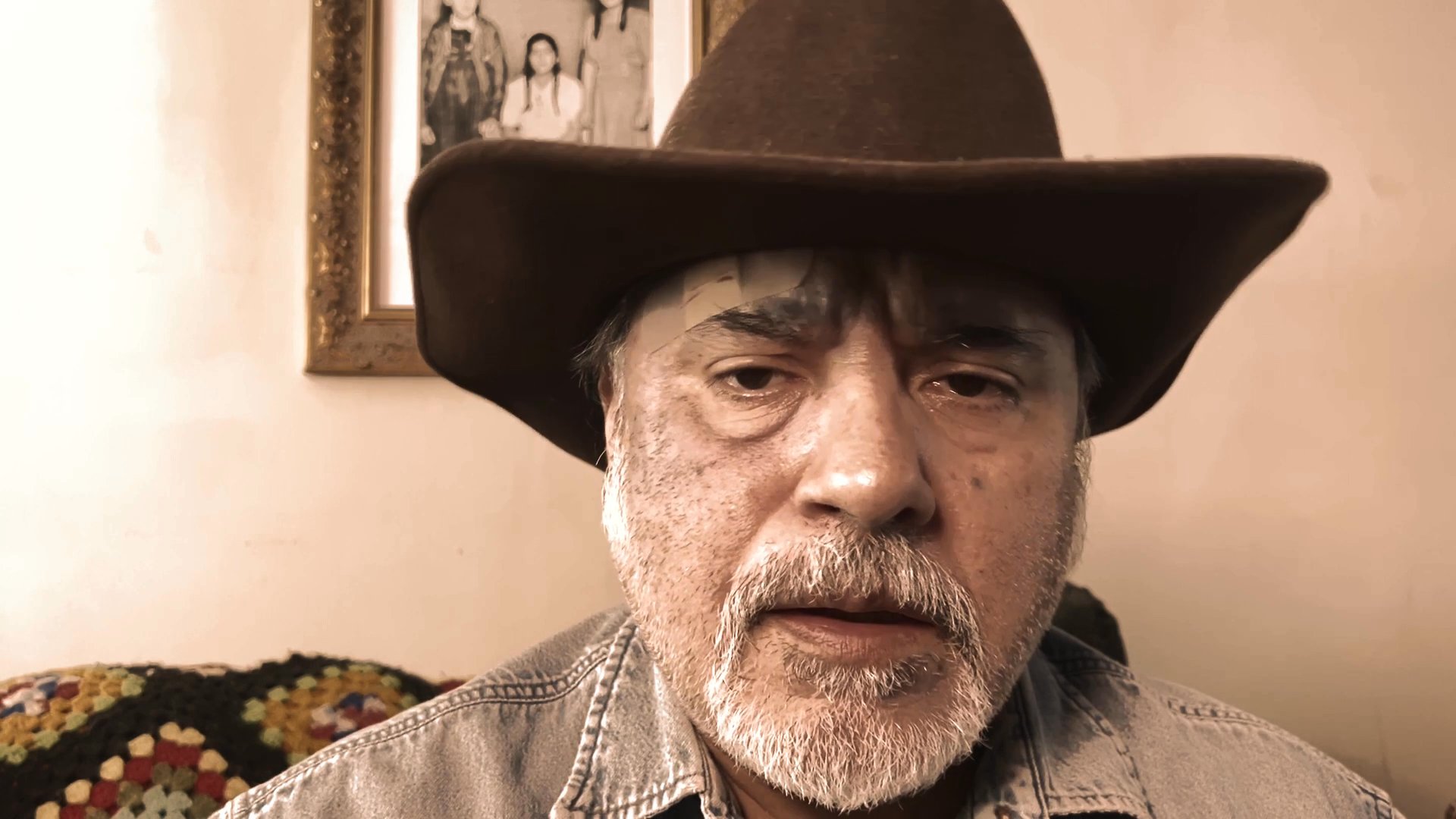 An older man wearing a cowboy hat delivers a serious message in an indoor setting, framed within a digital terminal interface.