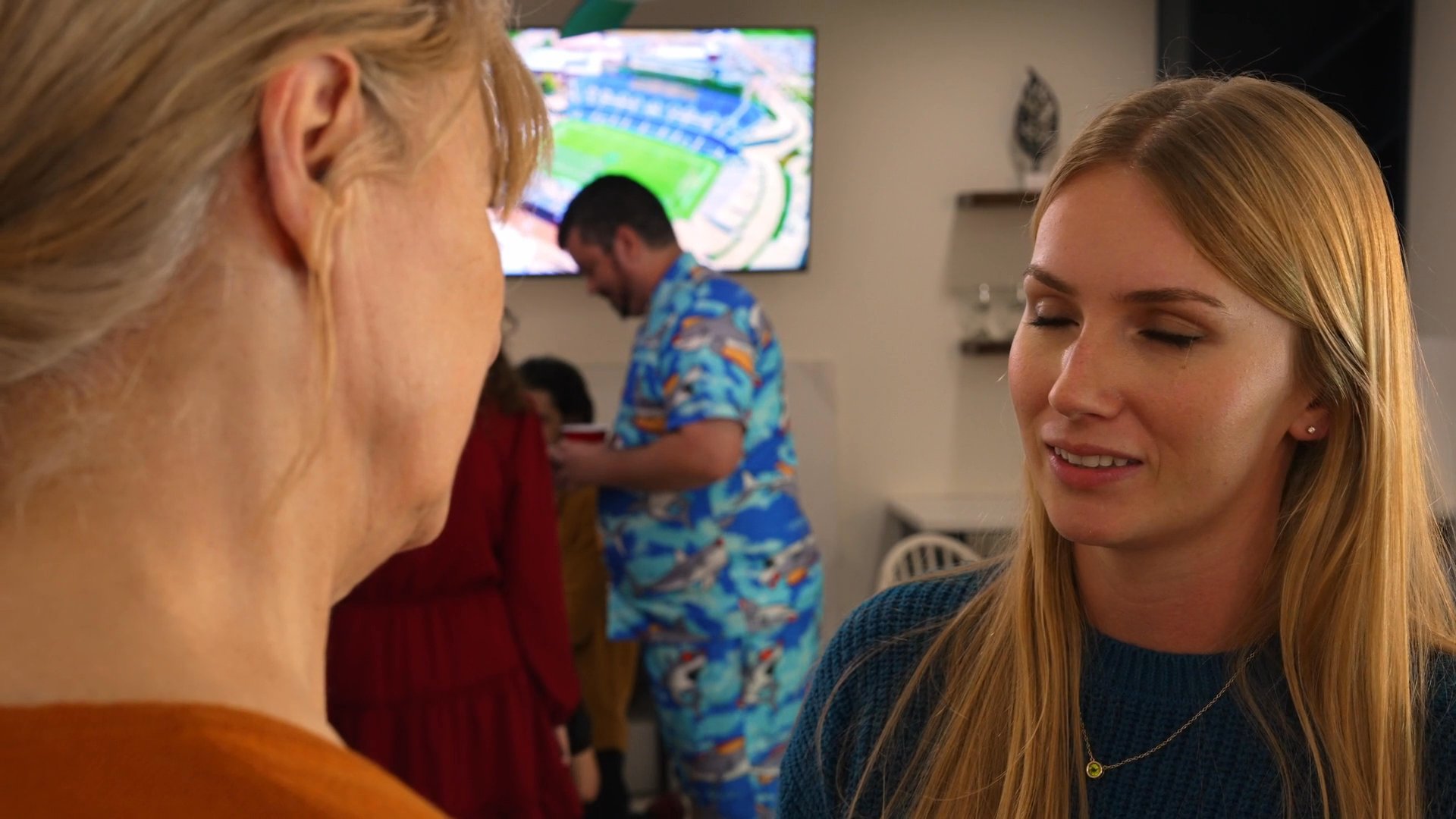 Two women engage in a tense, animated conversation during a social gathering in a domestic kitchen. The interaction culminates in a brief, assertive physical gesture.
