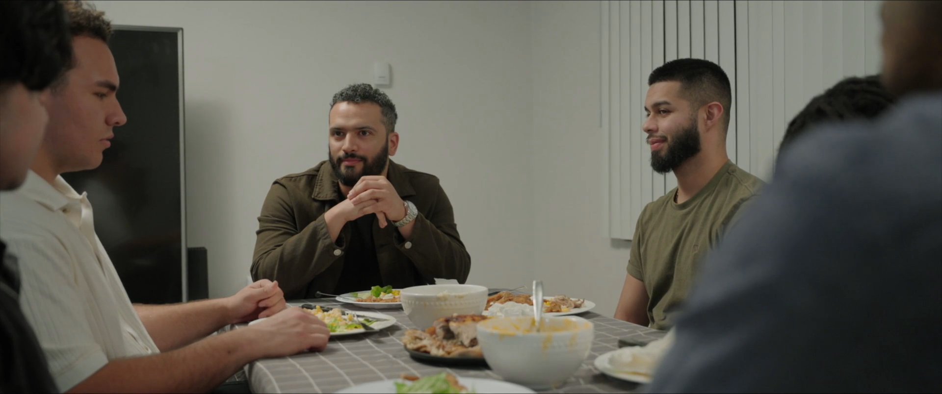 A group of friends engages in conversation while sharing a meal around a dining table. The interaction reflects a casual social gathering in a domestic setting.