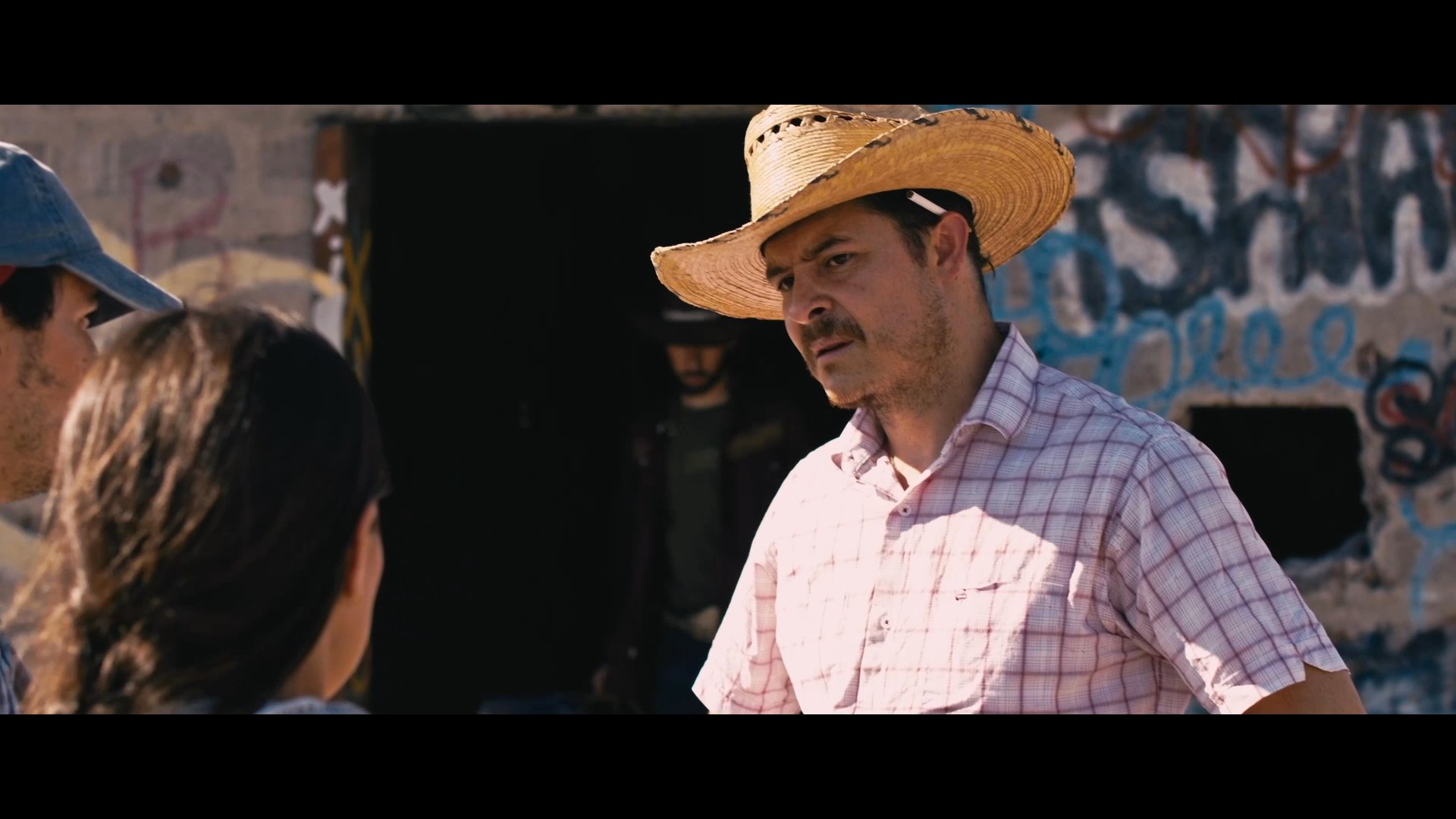 A man wearing a cowboy hat intimidates and coerces another individual into acting as a drug mule. The tense interaction takes place in a desert setting with another accomplice present.