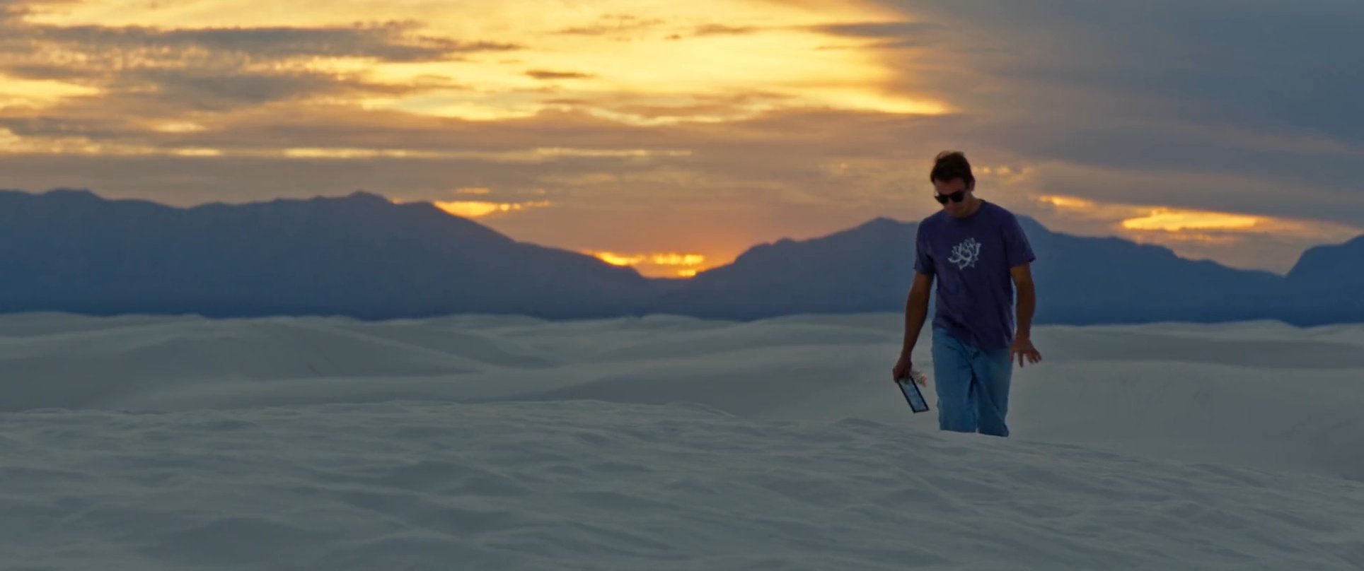 A man walks across white gypsum dunes toward the camera during a vibrant sunset with distant mountains in the background.