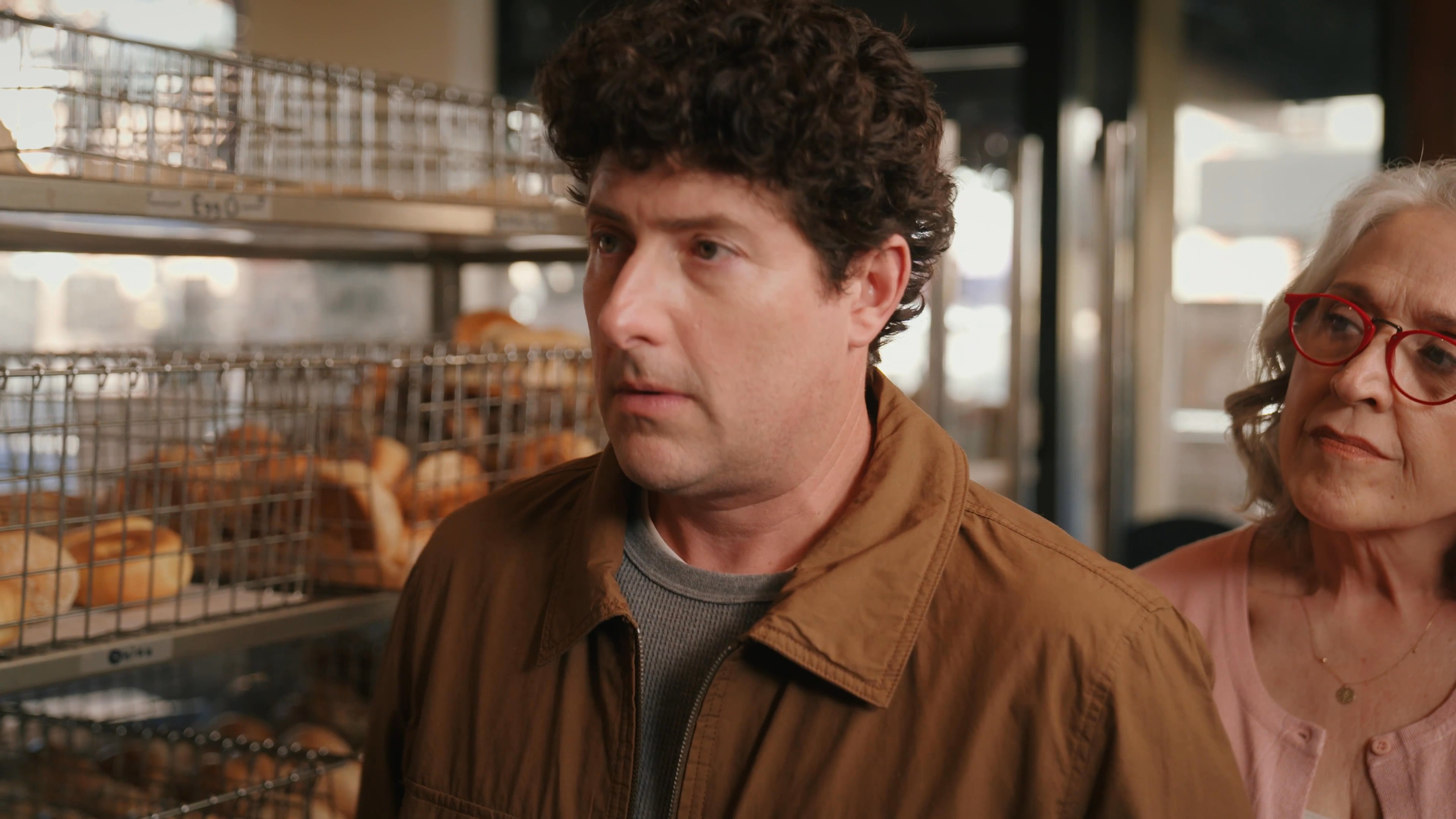 A man in a bakery receives a digital call from his mother while interacting with other customers. The scene captures a moment of modern technology disrupting a casual retail social setting.
