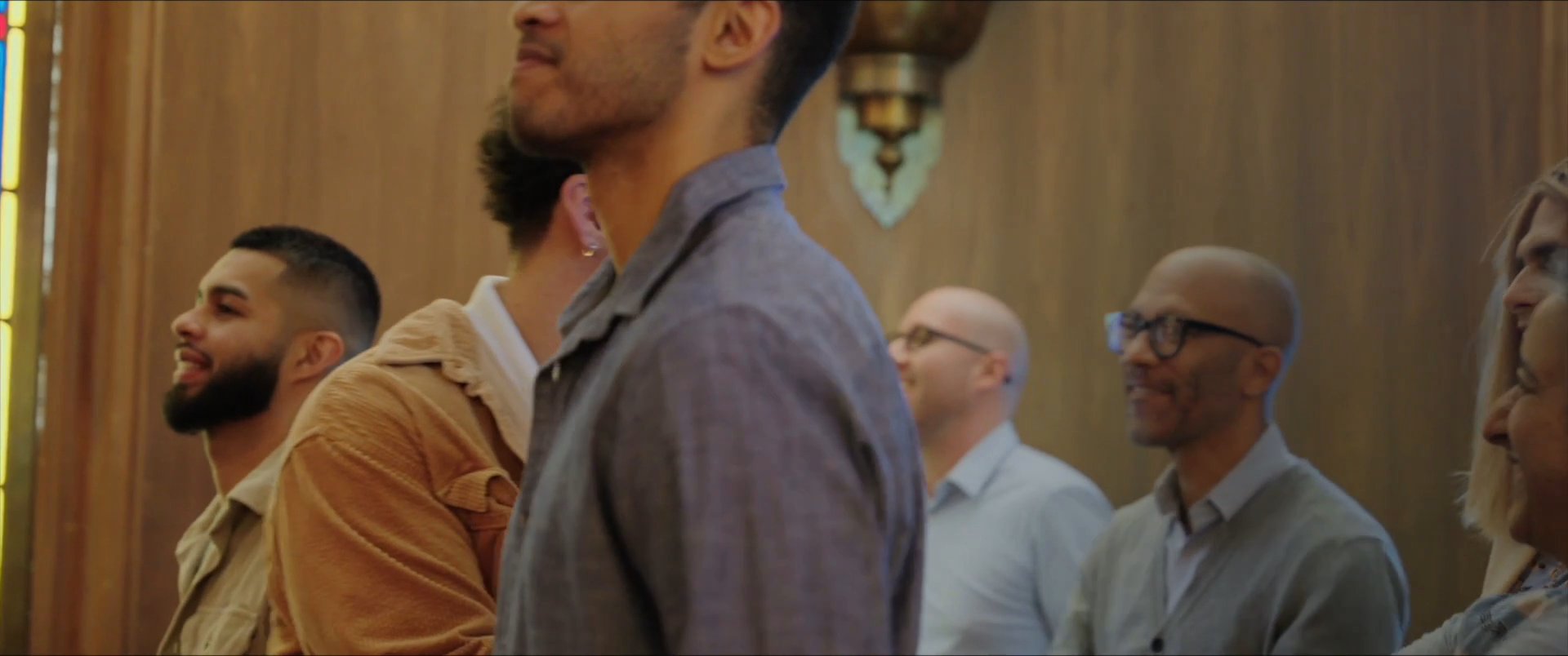 Diverse individuals engage in friendly conversation and interaction within a warm, wood-paneled interior space featuring stained glass accents. The atmosphere reflects a sense of community and shared presence during a social gathering.
