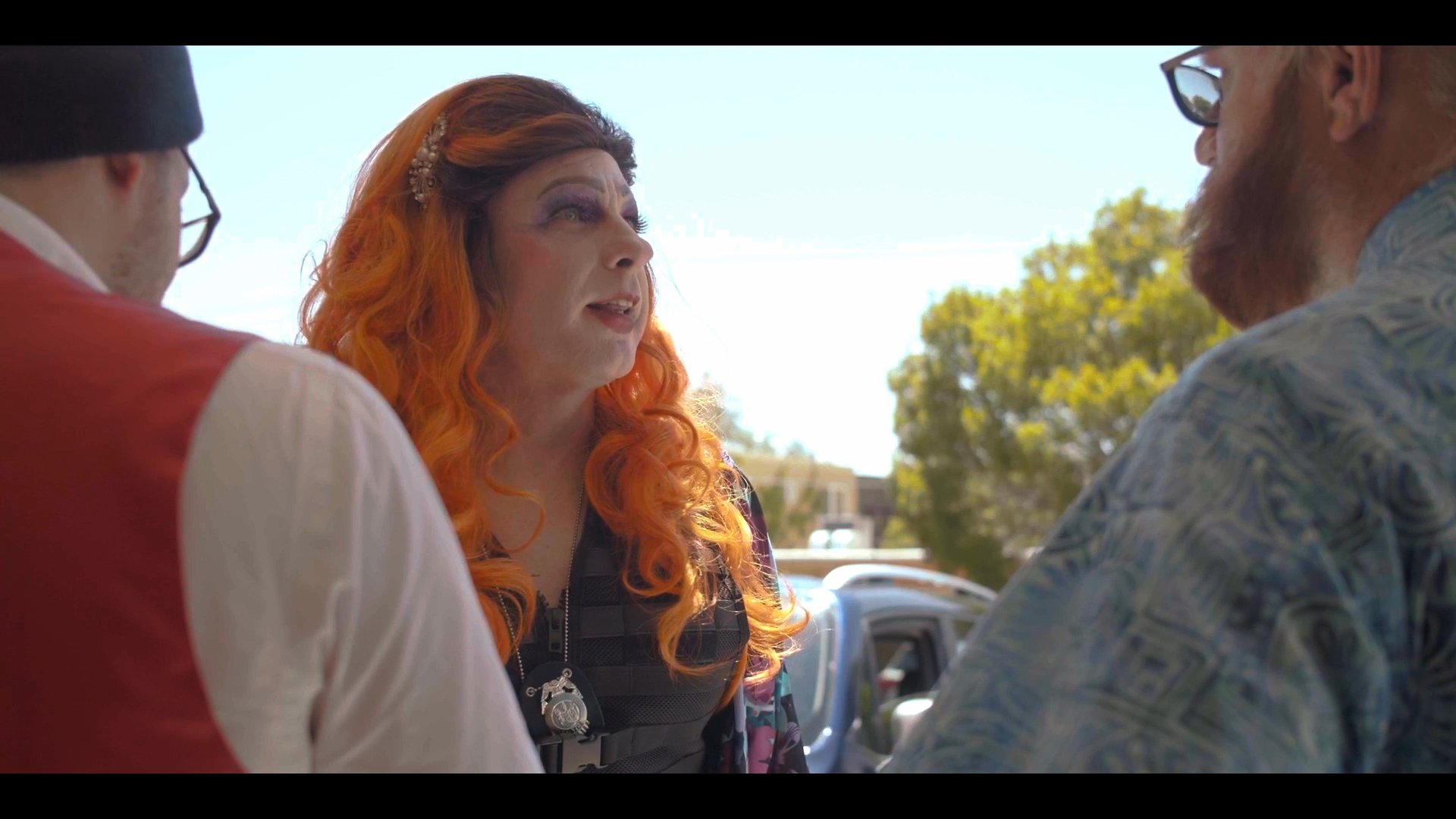 A group of individuals in distinct attire engages in an animated conversation in an outdoor setting. The interaction centers on a performer in elaborate makeup and a vibrant orange wig.