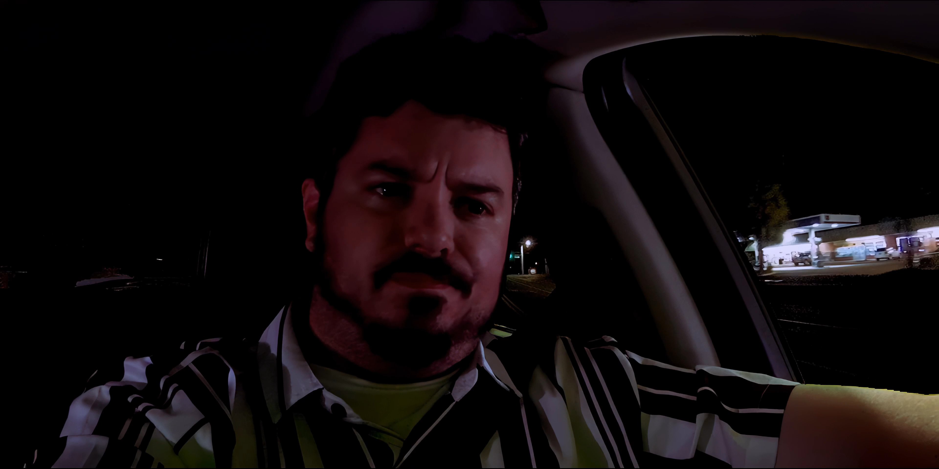 A man sits in the driver seat of a vehicle at night while engaged in a phone conversation. He periodically glances at his mounted smartphone throughout the ride.