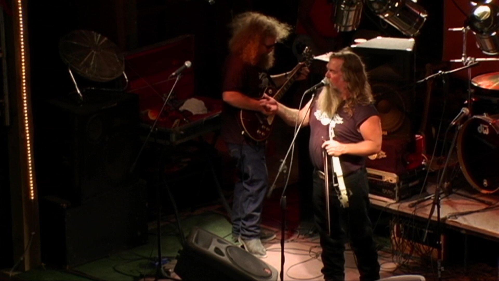 A long-haired, bearded lead singer performs on a concert stage with his band. The musician balances vocal duties with playing an acoustic instrument while other band members accompany him in a dimly lit venue.