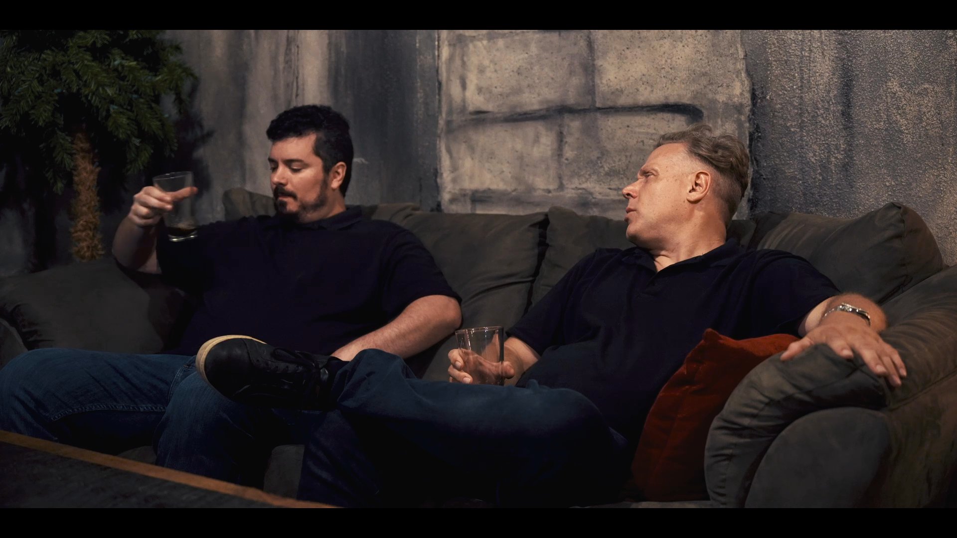Two men engage in a calm conversation while sitting on a sofa in a dimly lit room, punctuated by a brief, formal interaction with a newcomer at the end.