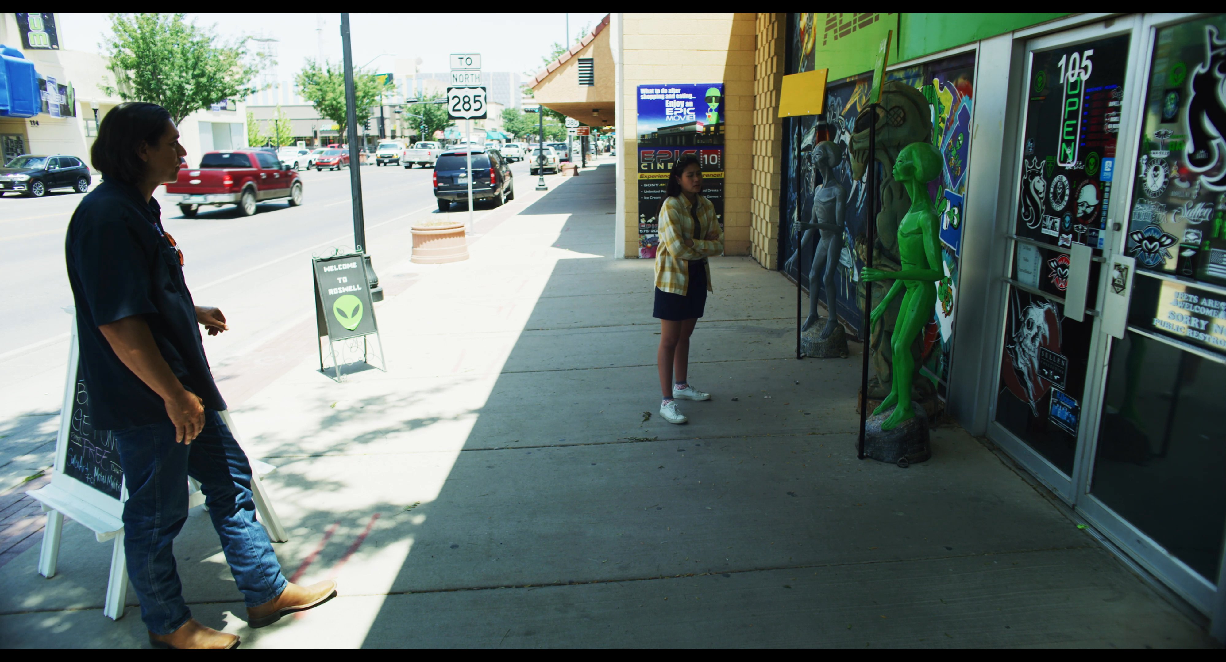 A woman stands in front of a themed alien storefront in Roswell before being approached by a man, leading to a brief conversation as they walk together along the sidewalk.