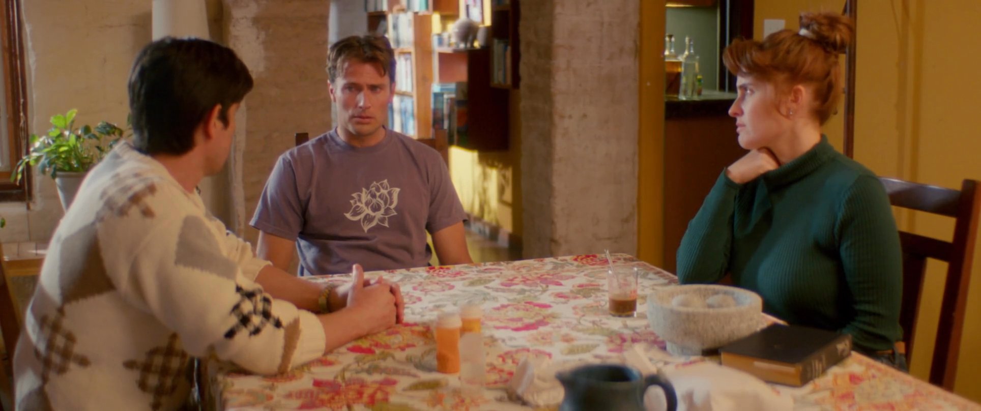 Three individuals sit around a dining table engaged in a serious and deliberate conversation within a residential setting. The interaction concludes as one participant stands up and exits the room.