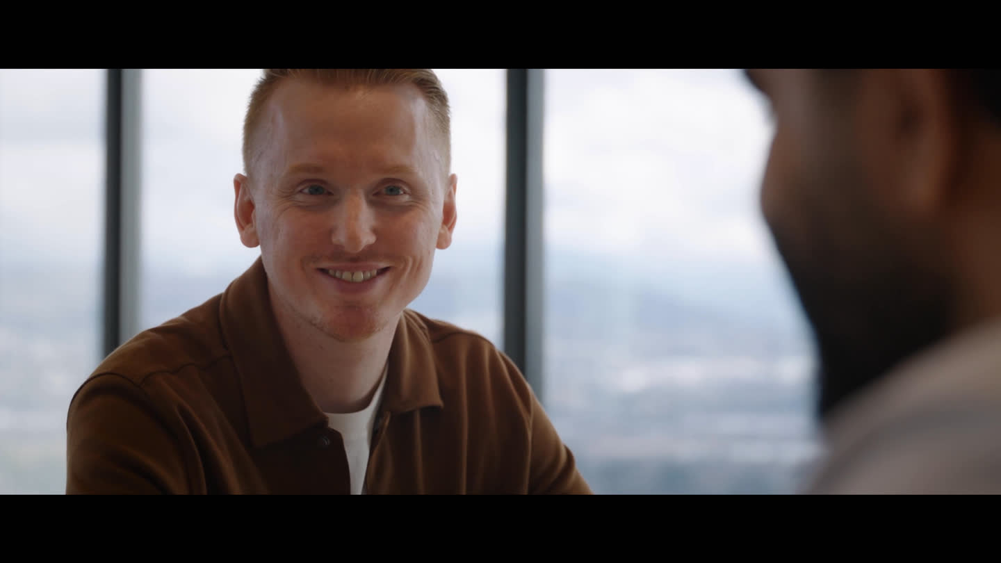 Film still: a smiling person in conversation beside an office window