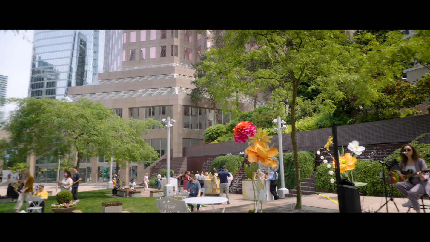 Film still: people gathered among event stalls and colourful decorations in a landscaped plaza
