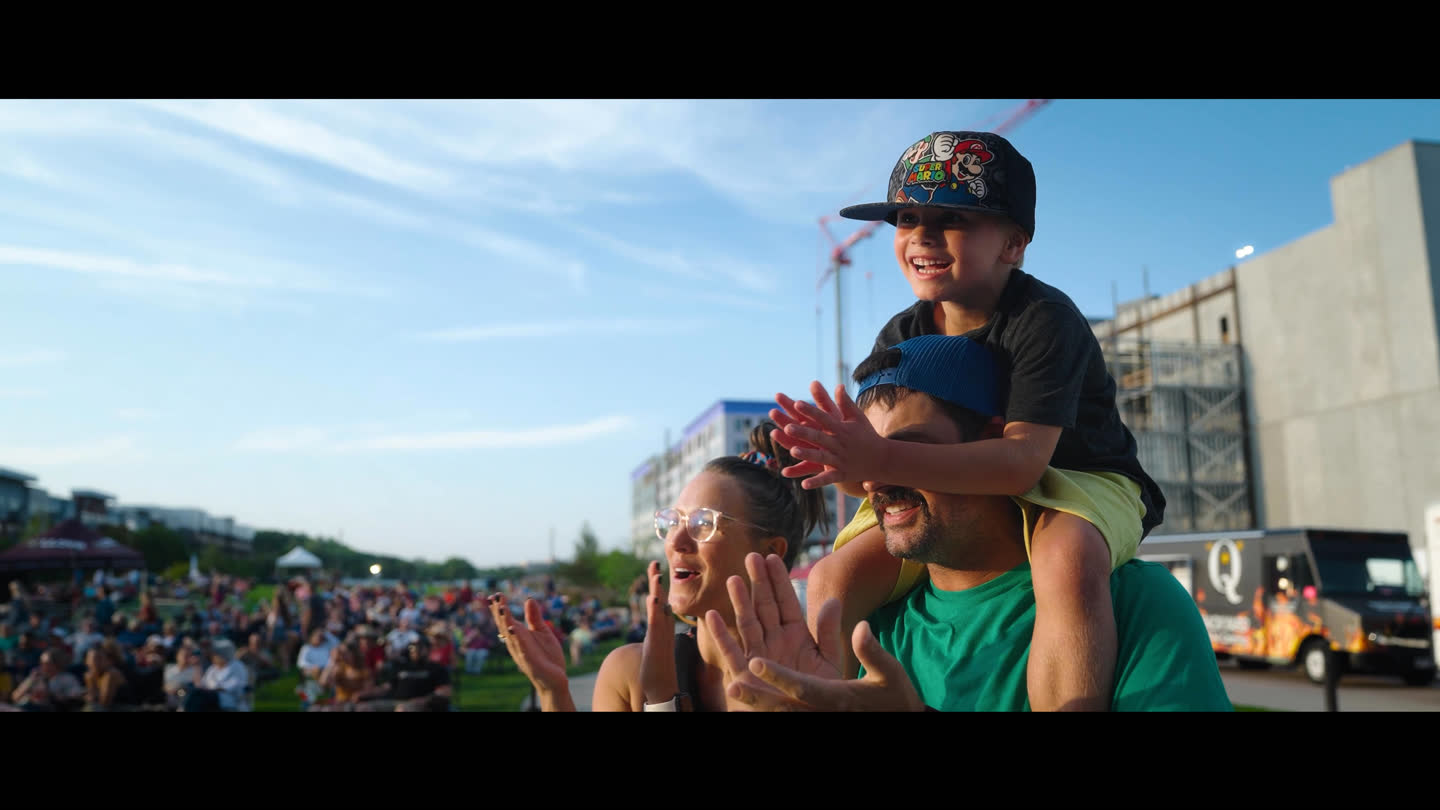 Film still: a smiling child on an adult’s shoulders in an outdoor event audience