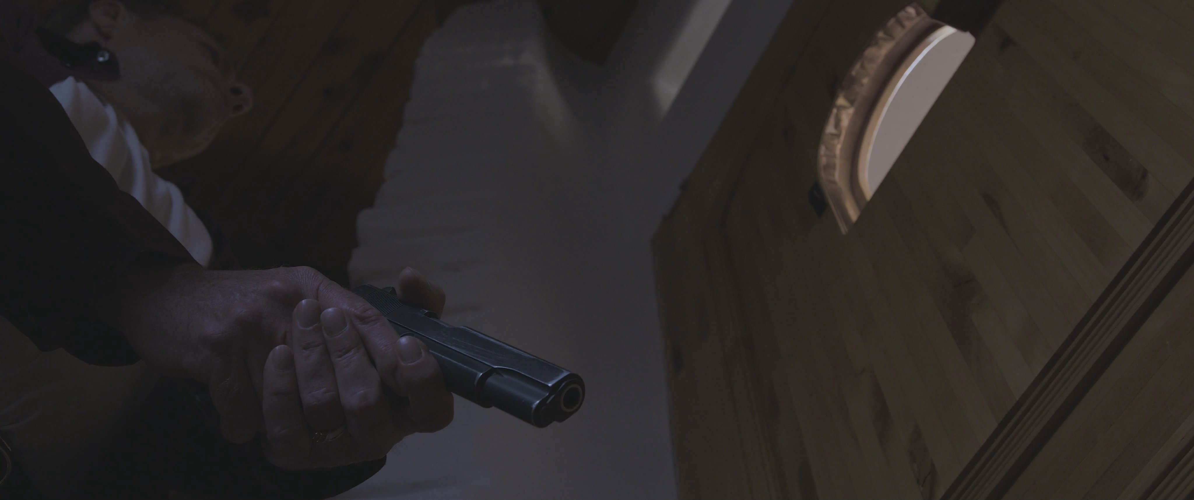 A man armed with a handgun monitors a woman through a small door aperture, creating a tense and threatening standoff.