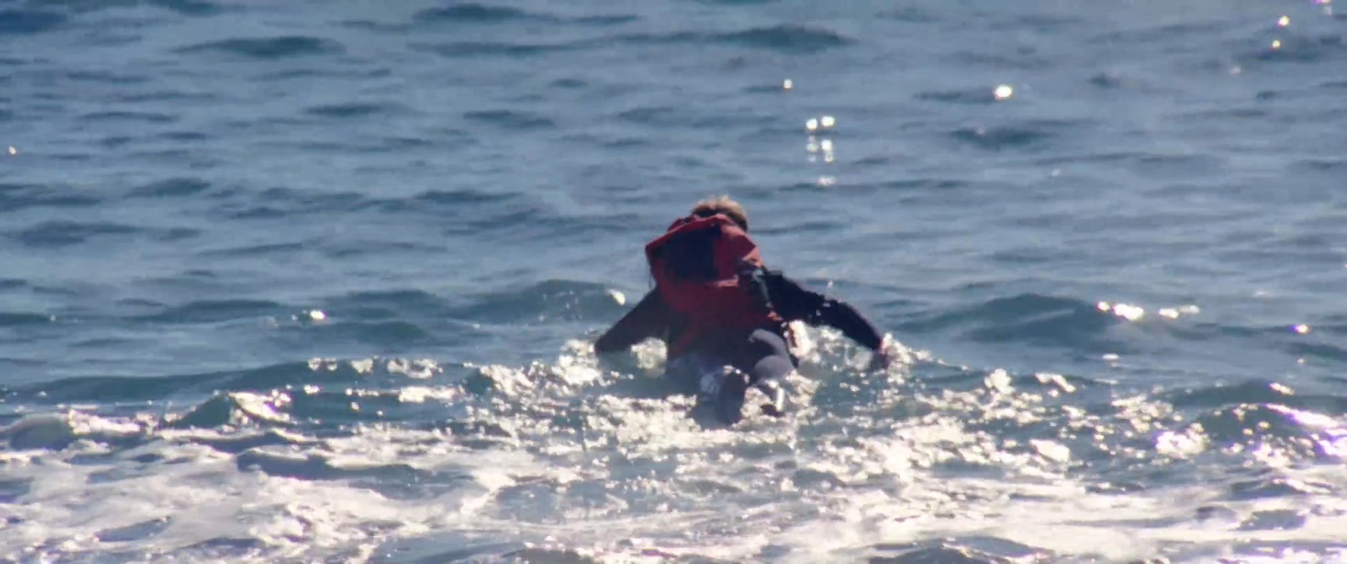 A surfer equipped with a wetsuit and life vest runs into the ocean to begin their session. The scene transitions to a close-up of a couple standing on the shore.