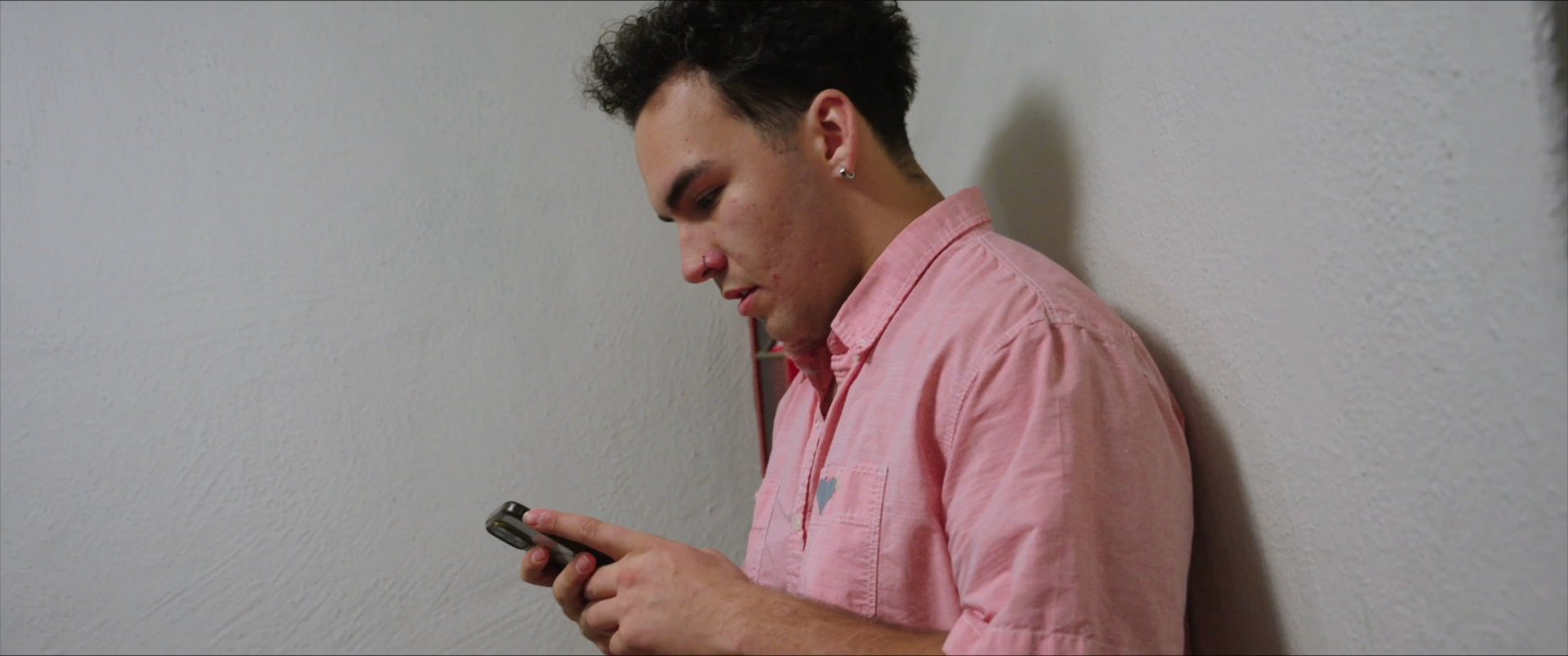A man in a pink shirt exhibits signs of stress and contemplation while standing in a stairwell. He retrieves his mobile phone to initiate a phone conversation.