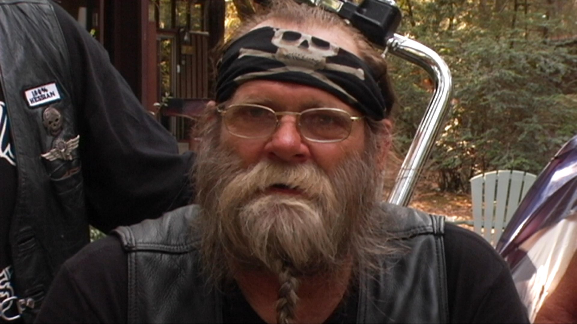 Various members of motorcycle clubs, including Van Camirillo and Sonny Barger, share perspectives in outdoor interview settings while a musician plays guitar.