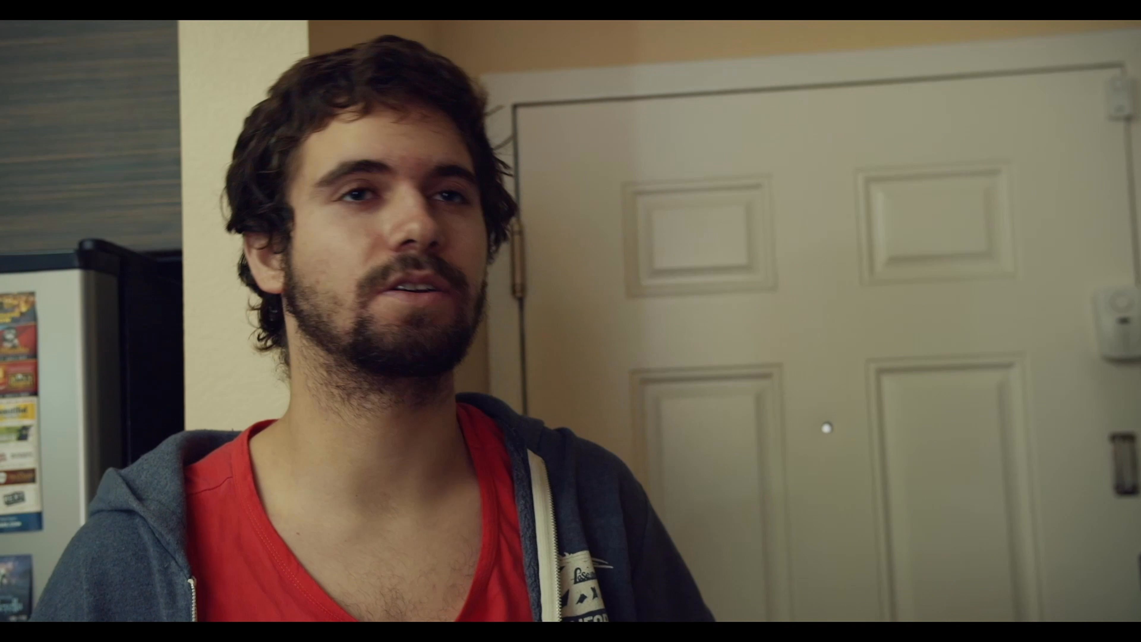 Three young men engage in a casual conversation within an apartment living space, characterized by relaxed interactions and expressive gestures.