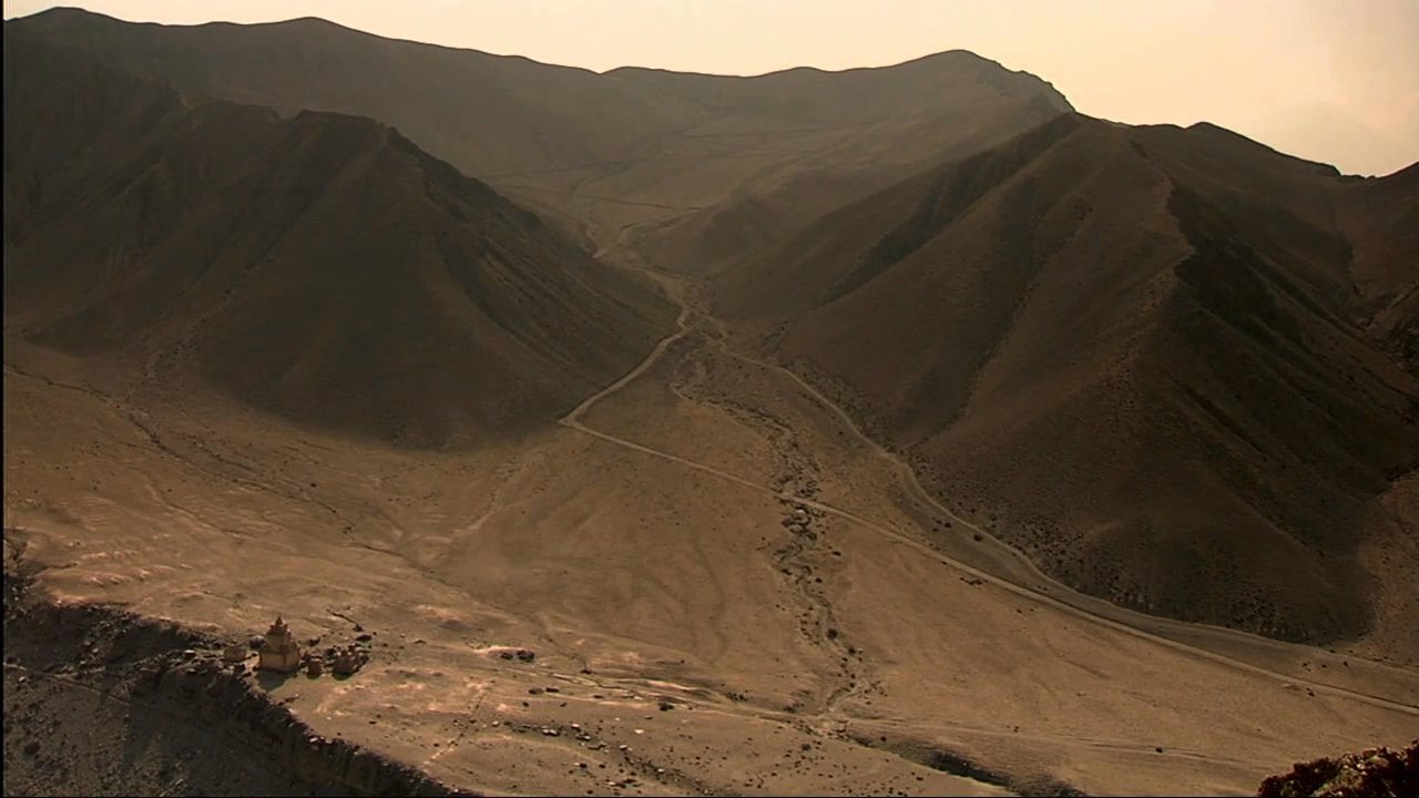 A narrator discusses regional heritage while showcasing aerial views of historic stupas set against rugged mountain terrain and local workers carrying fuel supplies. The footage highlights the intersection of ancient architectural traditions and daily life in high-altitude environments.