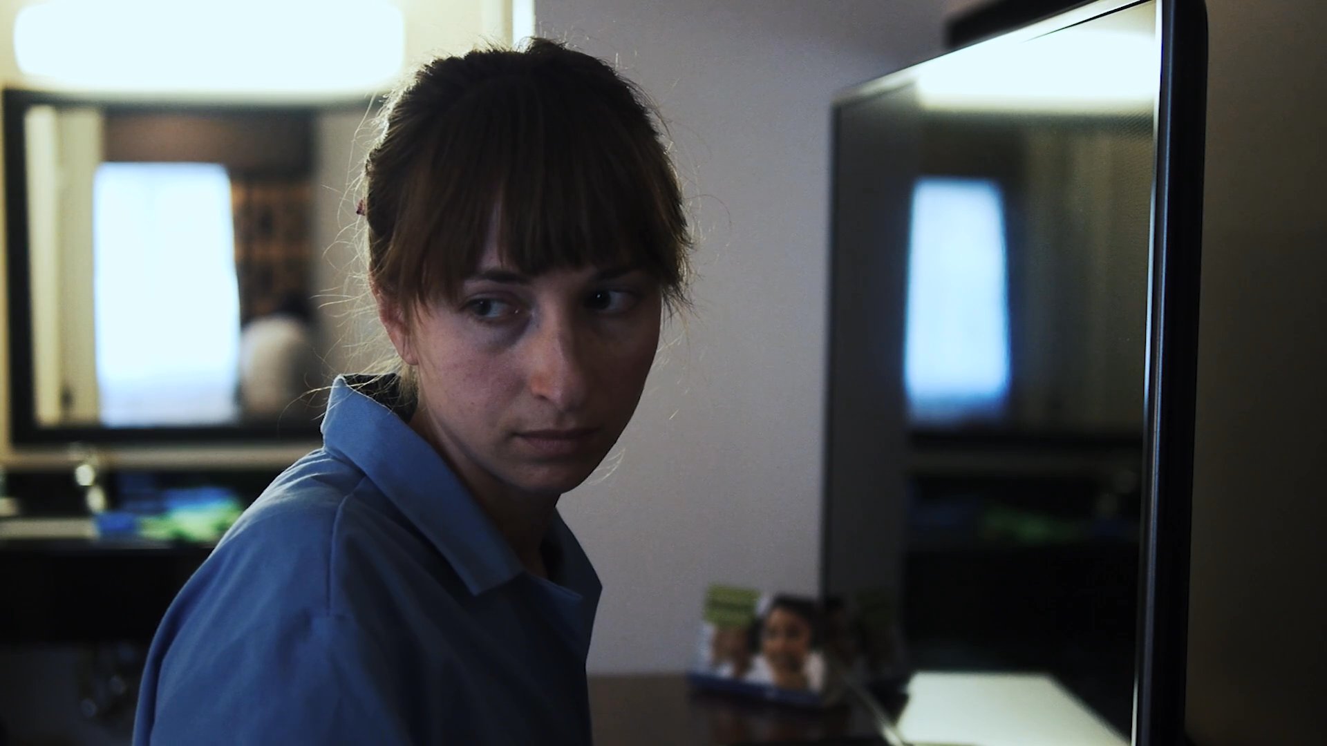 A man and a woman navigate a quiet, strained moment within the confines of a hotel room. Their measured movements and avoidant gazes emphasize a palpable sense of emotional distance and contemplation.