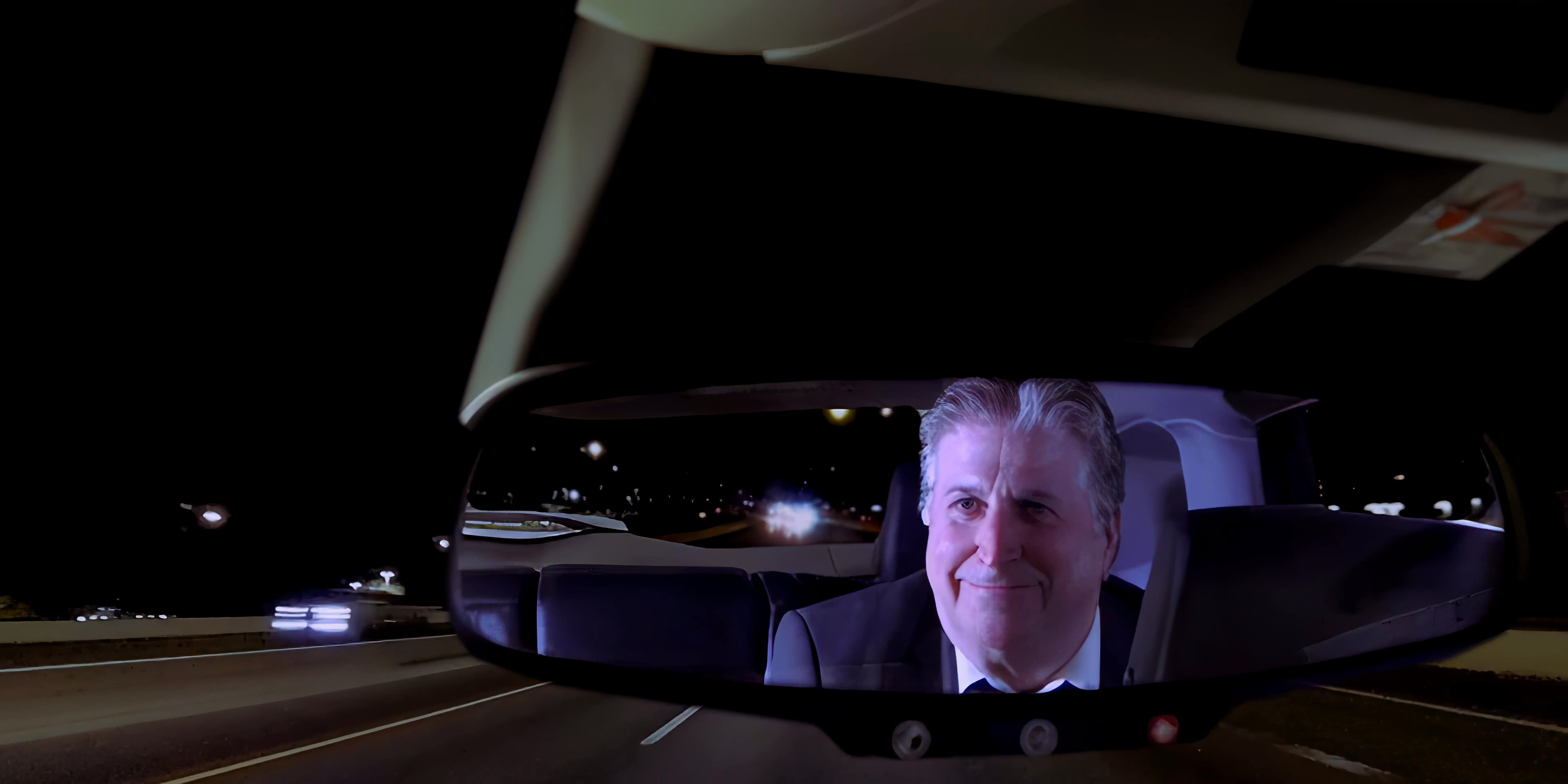 A man drives at night while participating in a video call displayed on his dashboard-mounted smartphone. The reflection of an older man appears in the rearview mirror as the driver maintains focus on the road.