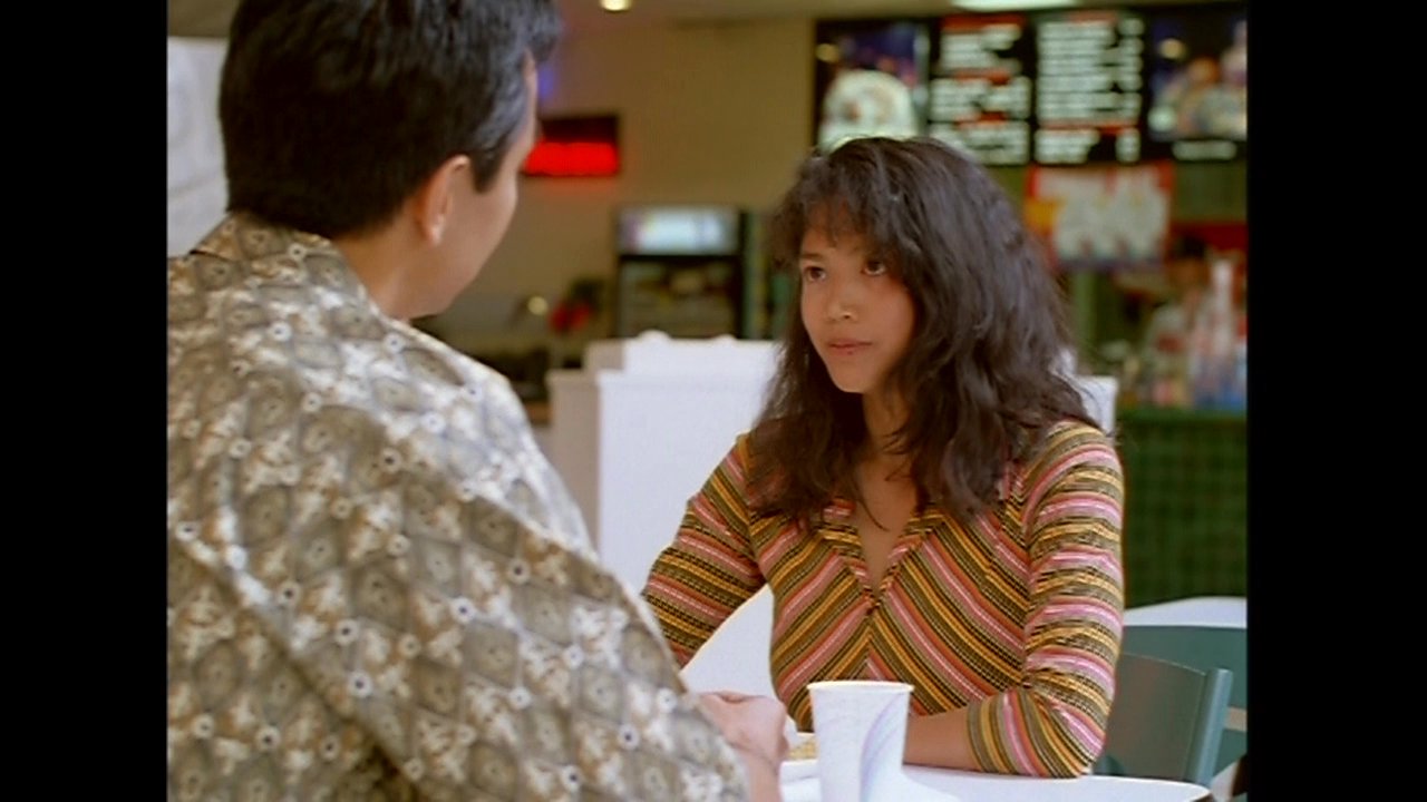 A man and a woman engage in a focused, seated conversation within a casual dining environment. The interaction maintains a serious tone as they exchange words across the table.