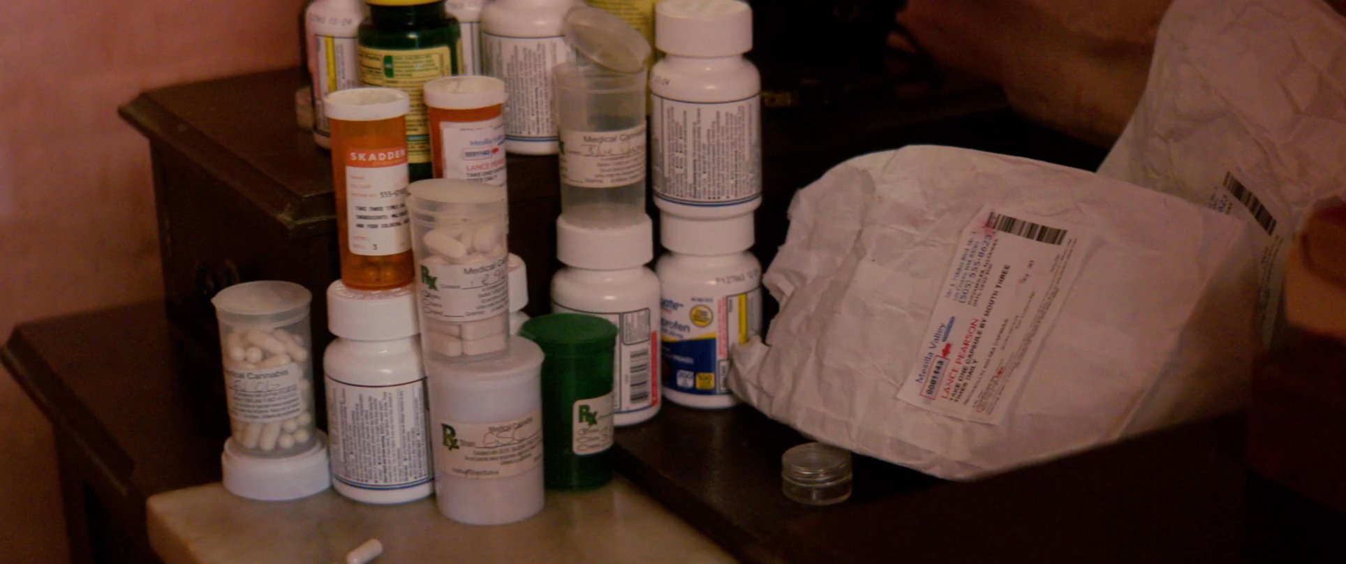 Two individuals engage in a focused conversation within a residential interior while a collection of prescription medication containers sits nearby.