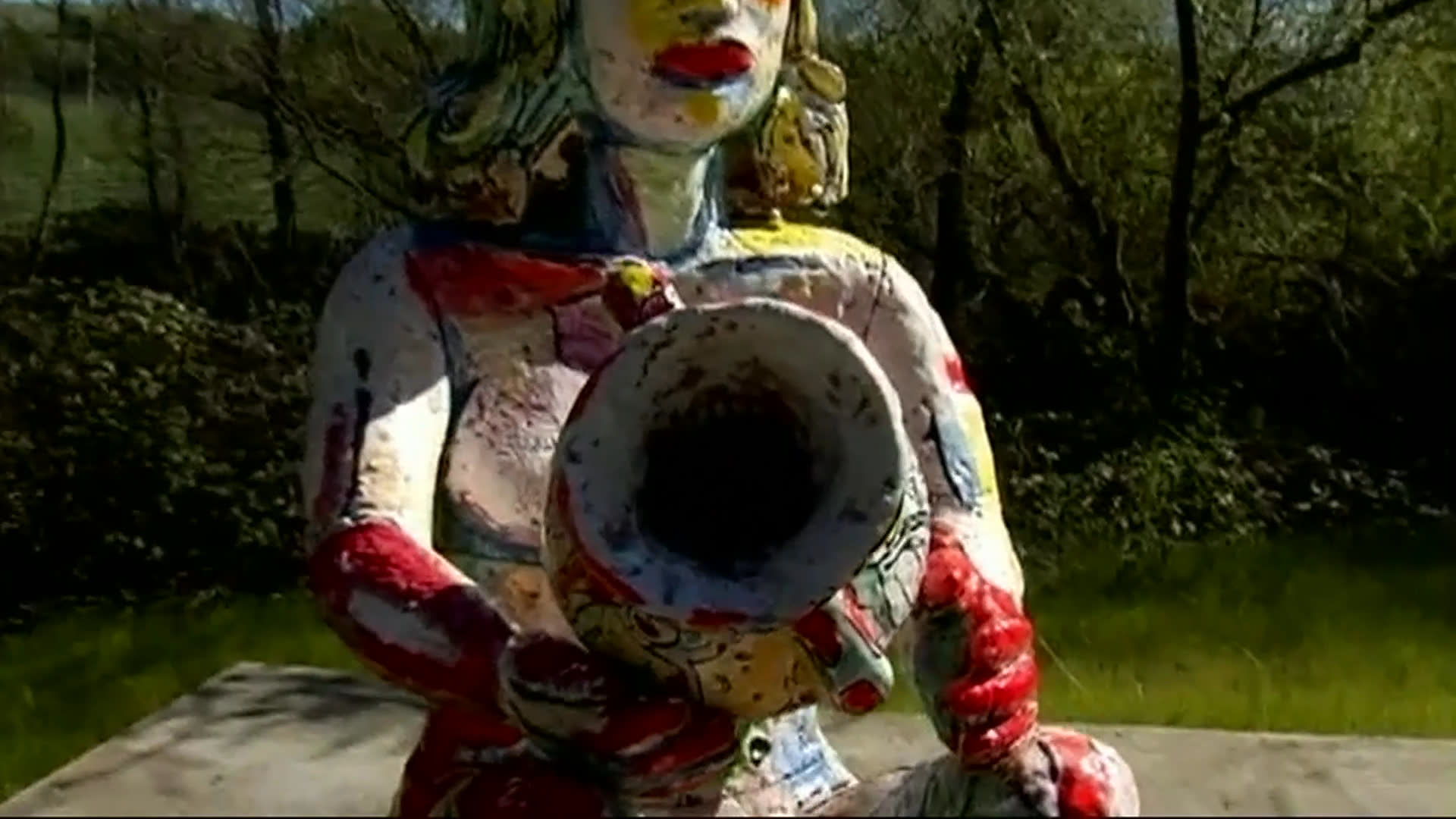 René di Rosa speaks outdoors in a garden, intercut with close views of a vividly painted human figure sculpture with a large circular opening. A rural field follows, with black drawn lines appearing across the landscape.