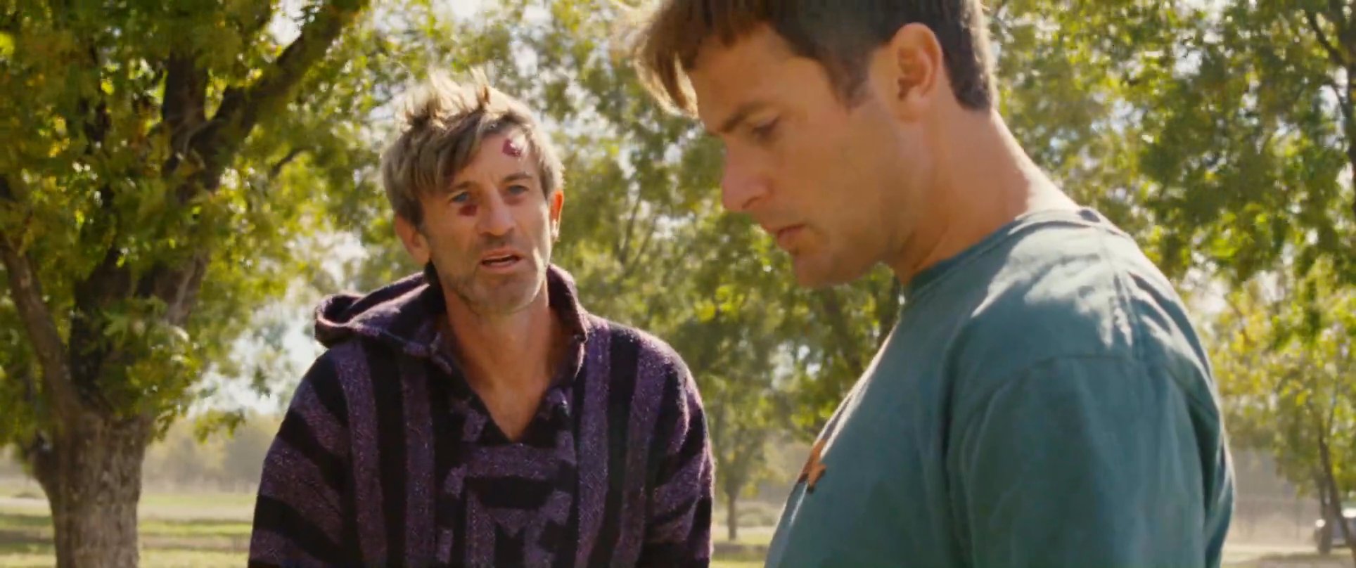 Two men stand in a sunlit orchard area engaging in a serious, quiet conversation. One individual gestures while speaking, while the other listens intently.