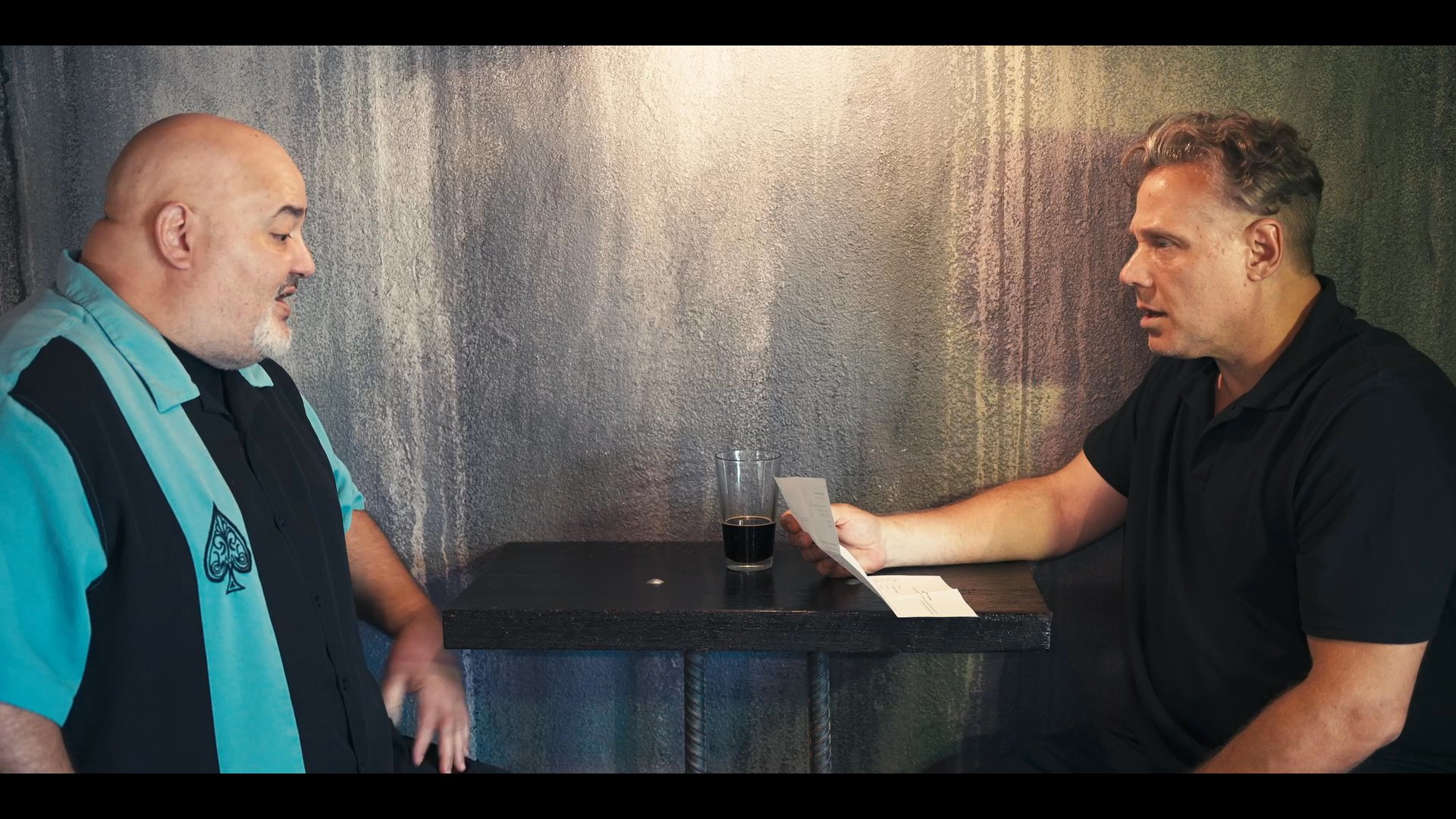 Two men sit across from each other at a table in a dimly lit room, engaged in a serious discussion while reviewing a document. Their focused body language suggests a formal consultation or negotiation process.