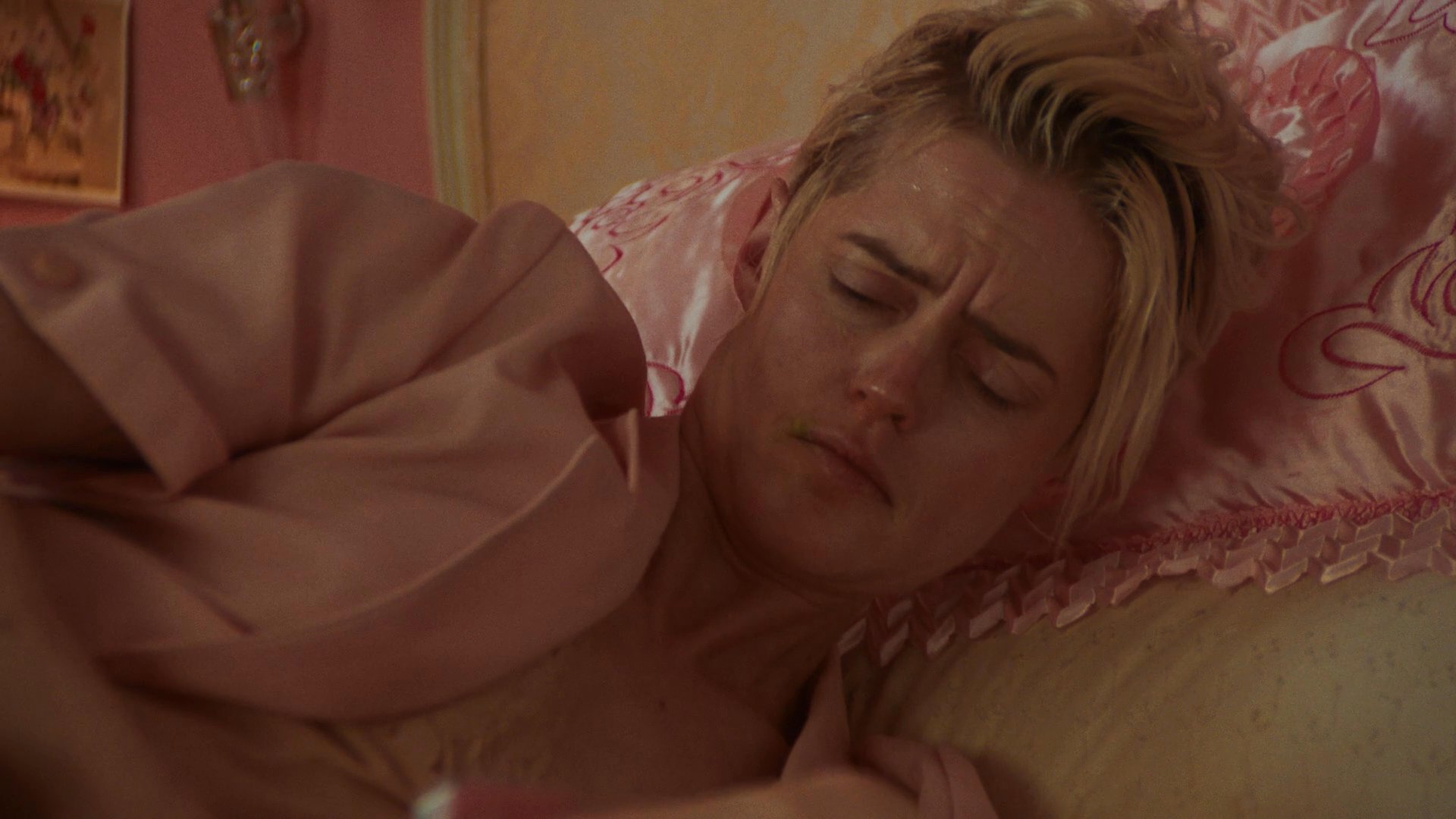 A woman lies on a pink-themed bed in a state of repose and reflection, surrounded by personal items.