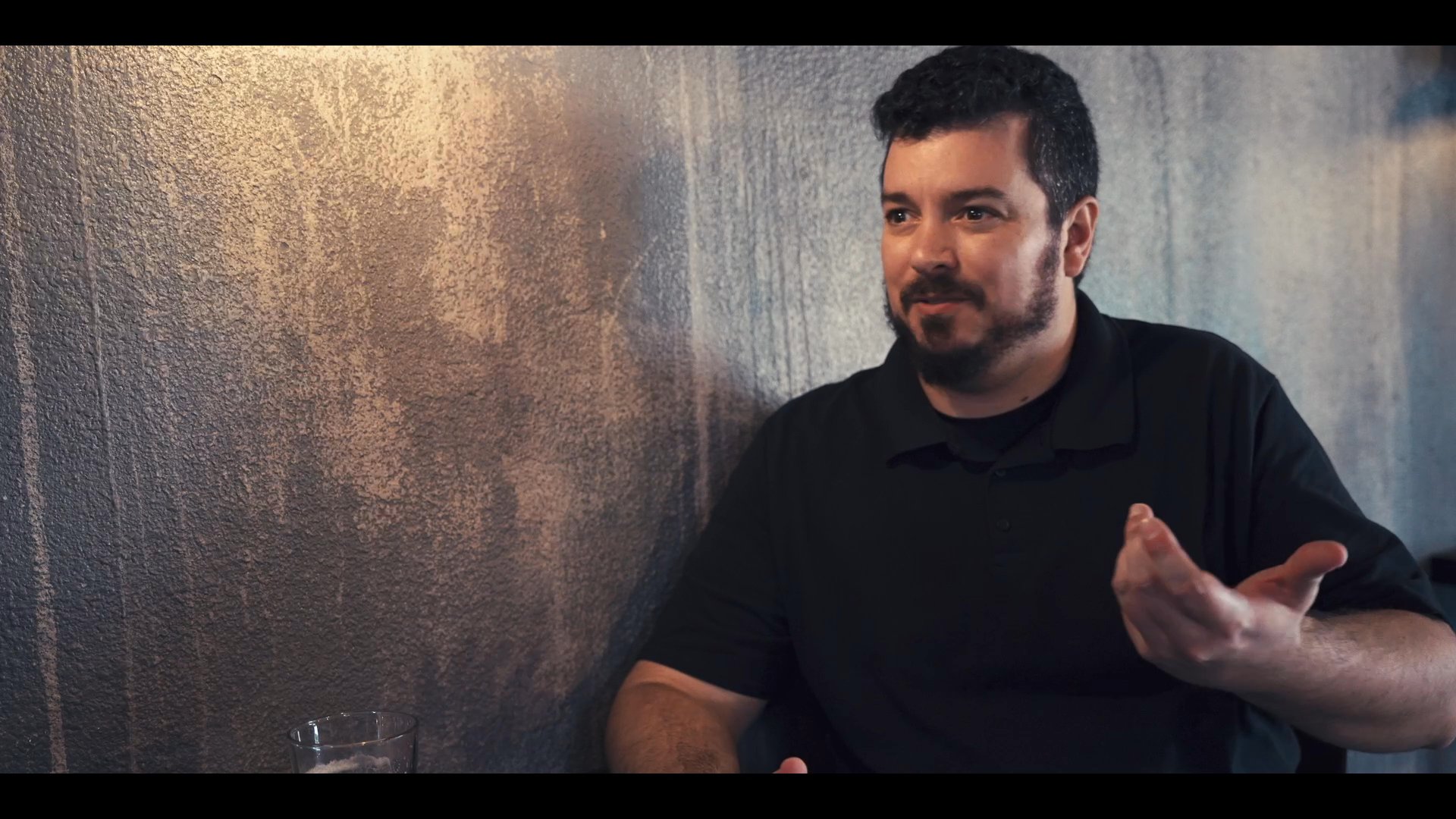 Two men sit at a table engaged in an active conversation, sharing perspectives in a casual setting. They alternate between speaking and listening as the discussion progresses.