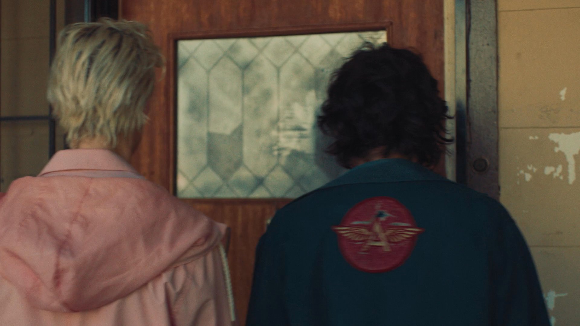 Two individuals approach a residential building and investigate the front entryway. One partner leans in to signal their arrival at the door.
