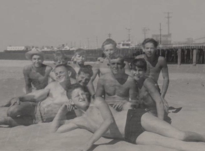 A man reminisces while looking at archival footage of a group of young men relaxing and swimming near a pier. The sequence transitions from a static group portrait on the sand to scenes of the individuals enjoying the ocean water.