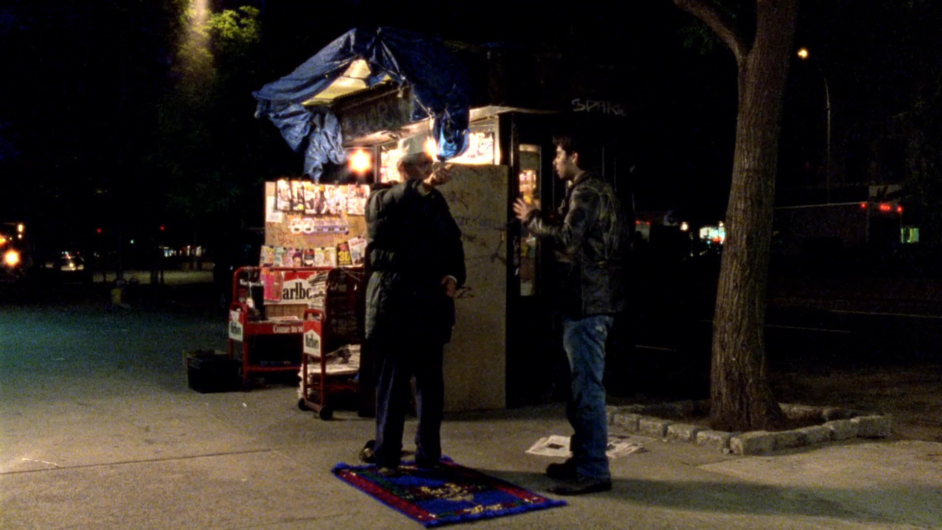 A man engages in a tense conversation with a newsstand vendor on a dimly lit urban sidewalk at night.