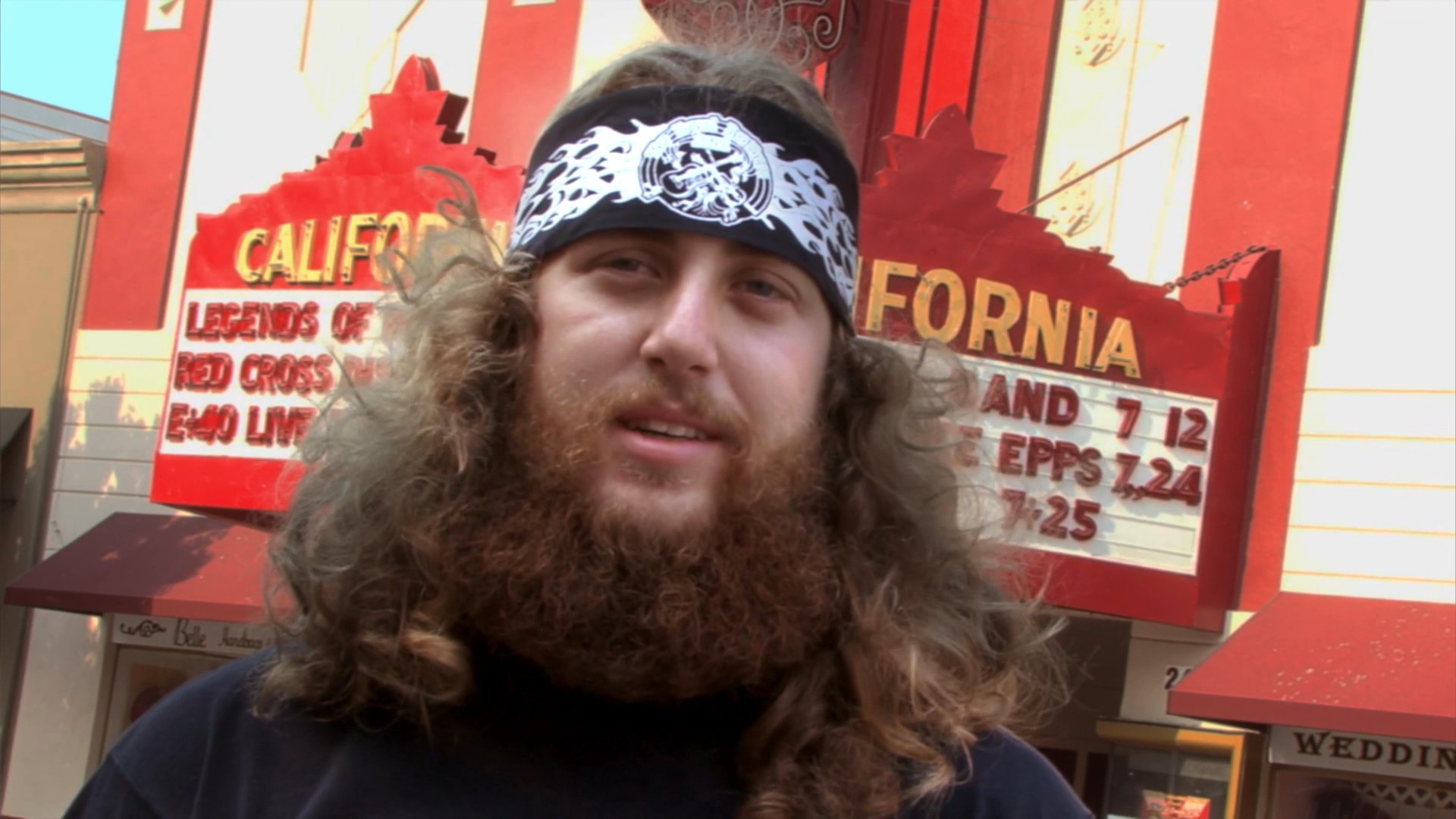 A man with long hair and a bandana speaks in front of the Fox Theater in downtown California, followed by a wide-angle view of the surrounding city street.