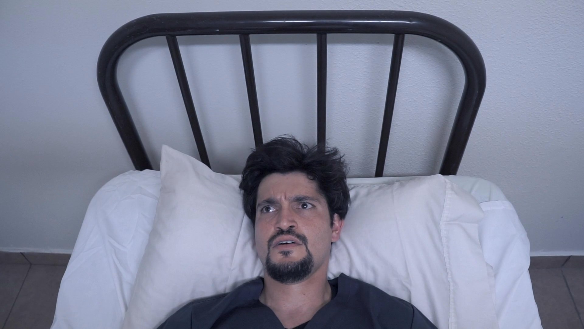 A man lies in a hospital-style bed, appearing distressed and restless as he experiences fragmented, disorienting visions. His emotional state transitions from contemplative to increasingly agitated within the confines of the room.