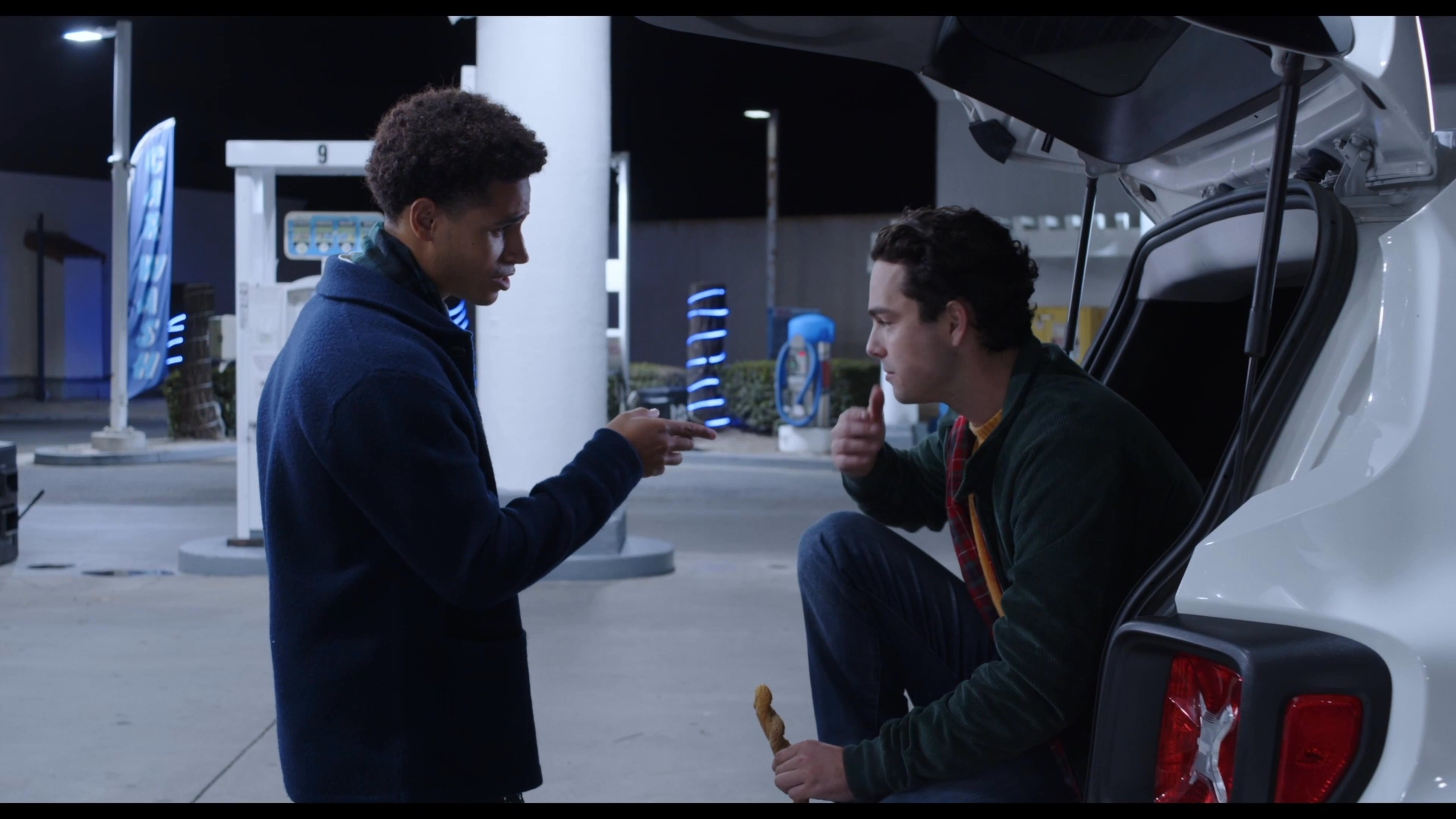 Two young men engage in a calm conversation while standing near an open vehicle trunk at a gas station at night.