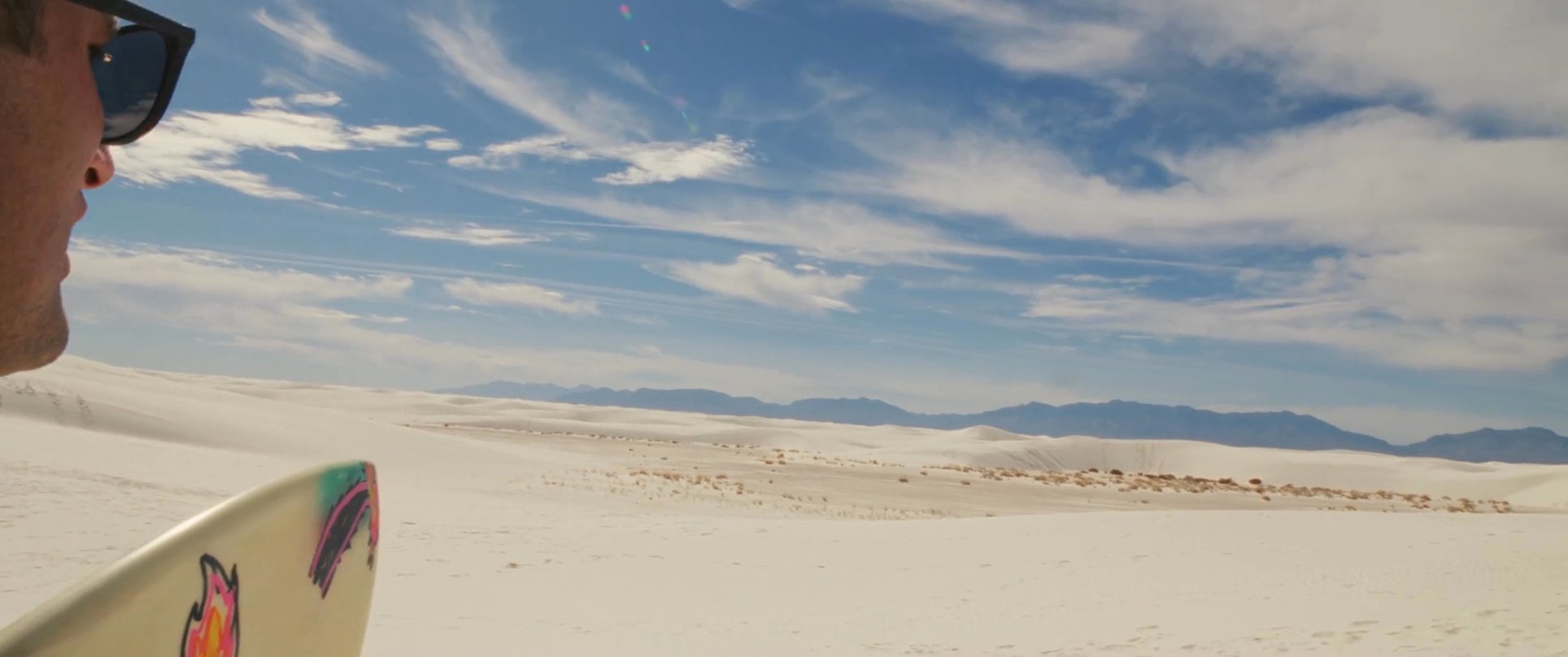 Two men carry surfboards across a vast, expansive white desert landscape under a clear blue sky. They observe their surroundings before continuing their journey through the dunes.