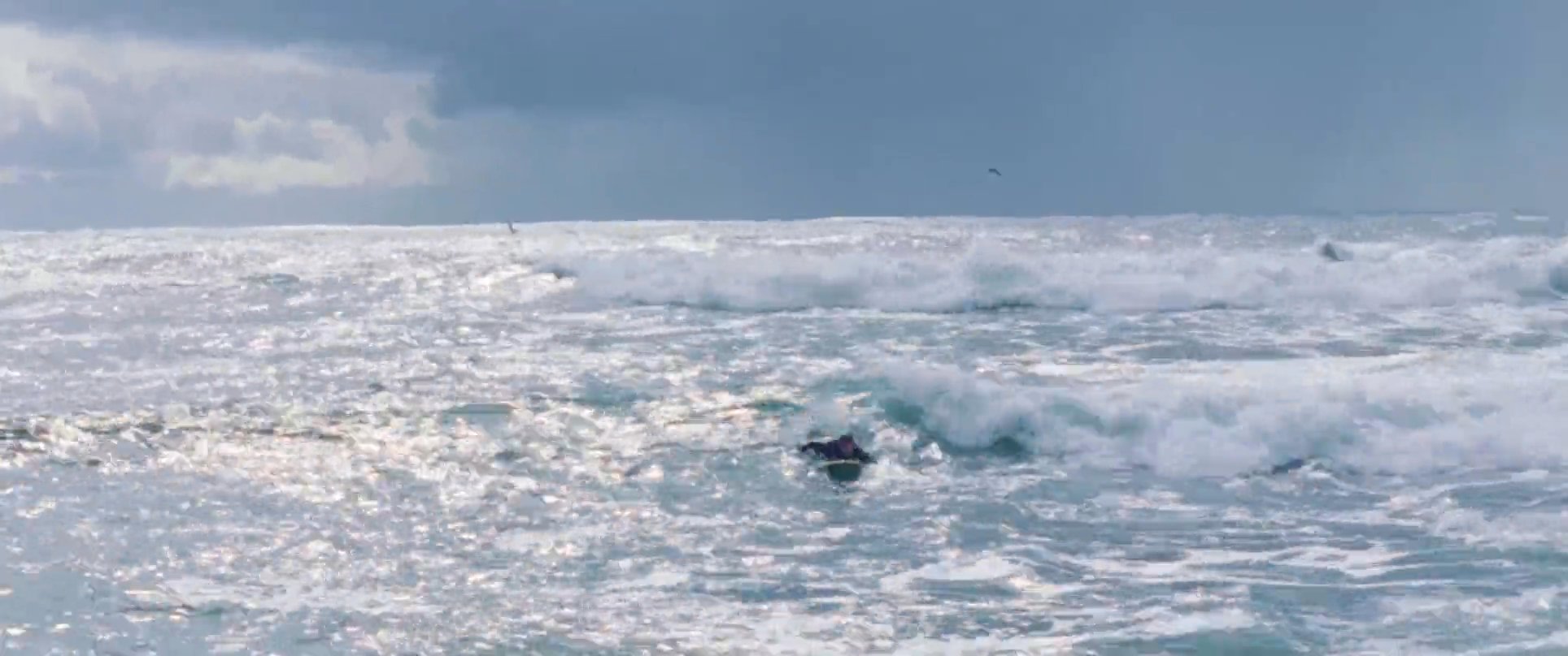 A surfer navigates open waters and catches a wave before wading toward the coastline in a wetsuit.