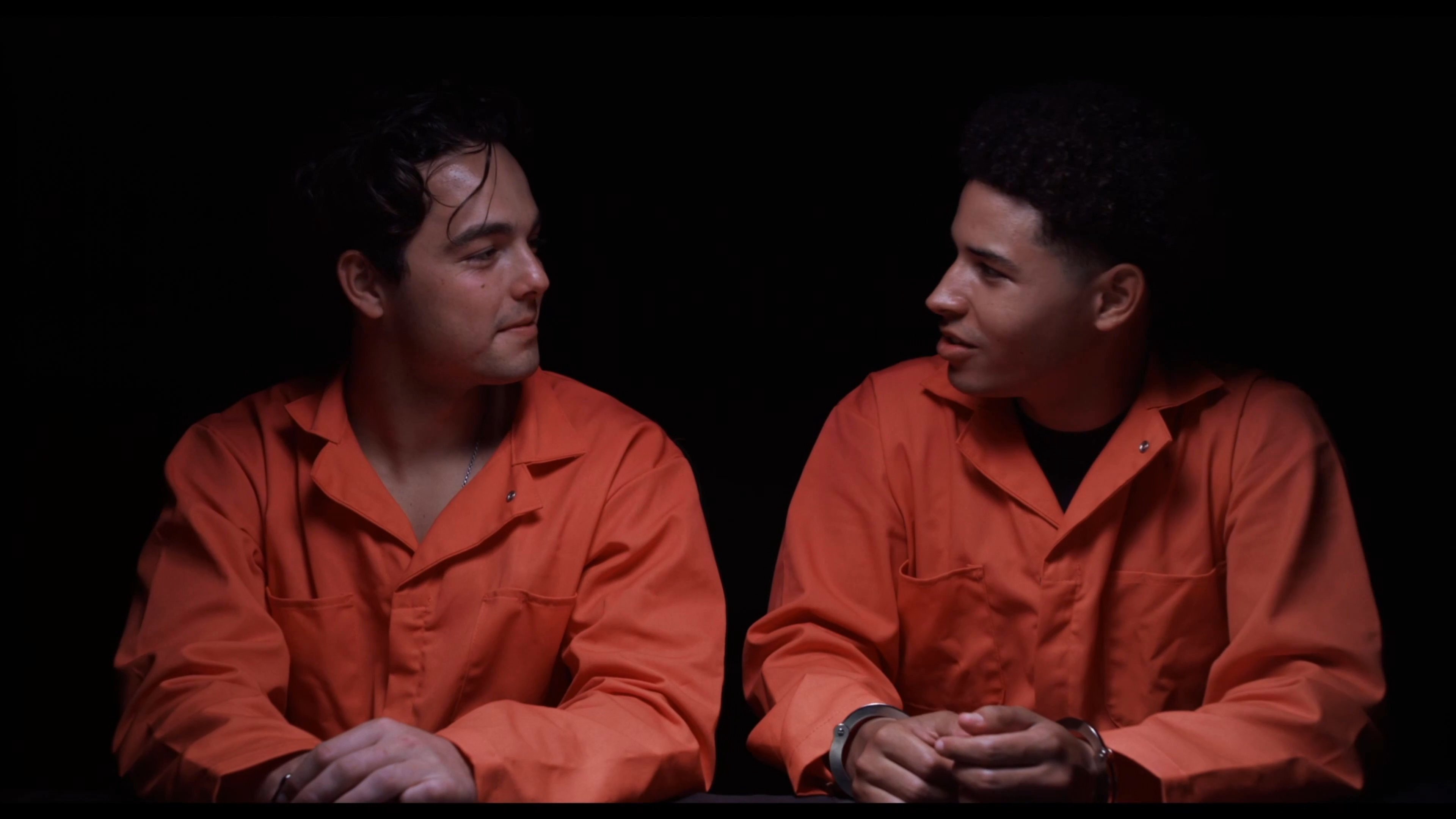 Two individuals dressed in orange prison jumpsuits sit in a dark, dimly lit environment, engaged in an intimate and focused conversation. The scene captures a moment of intense interaction between the two figures.