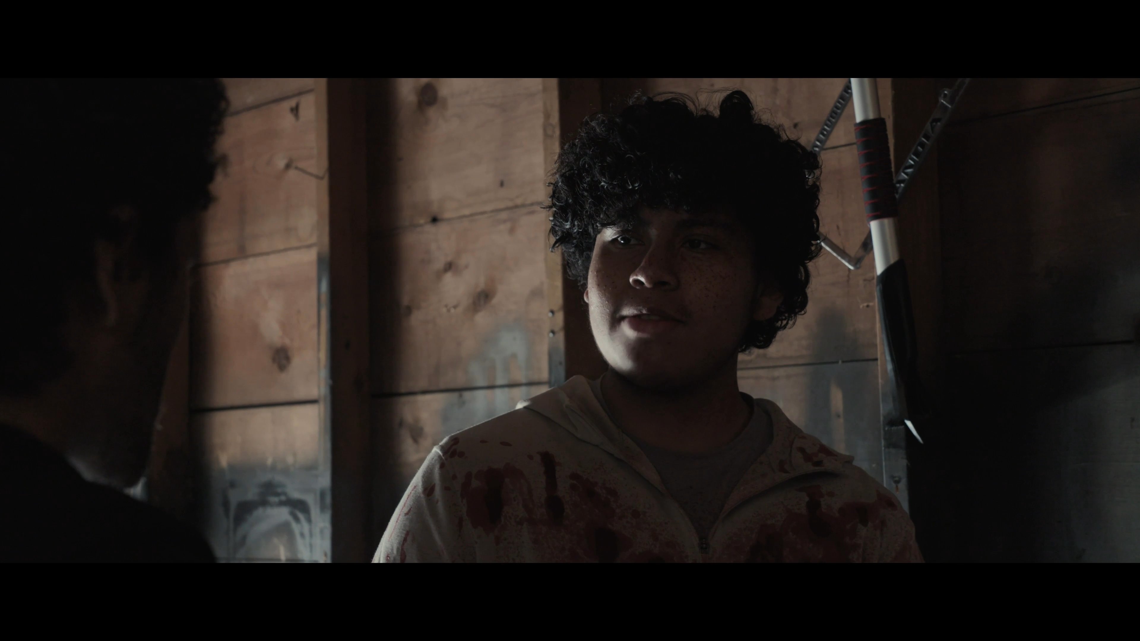 Two individuals engage in a serious, emotionally charged conversation within a dimly lit, rustic interior setting. The exchange maintains a persistent sense of urgency and interpersonal tension.