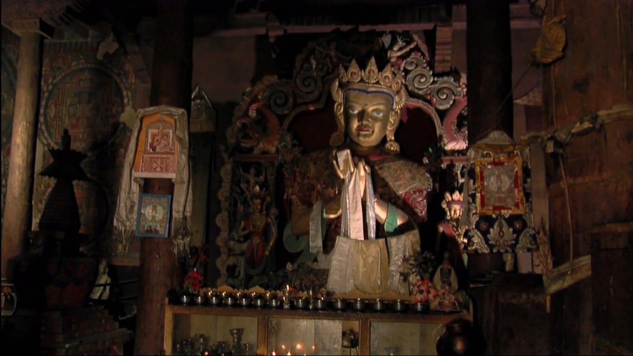 A monk in traditional attire performs a ritual dance for an audience before shifting to a discussion about spiritual practices within a Tibetan monastery. The footage highlights ornate religious statues and intricate temple iconography.