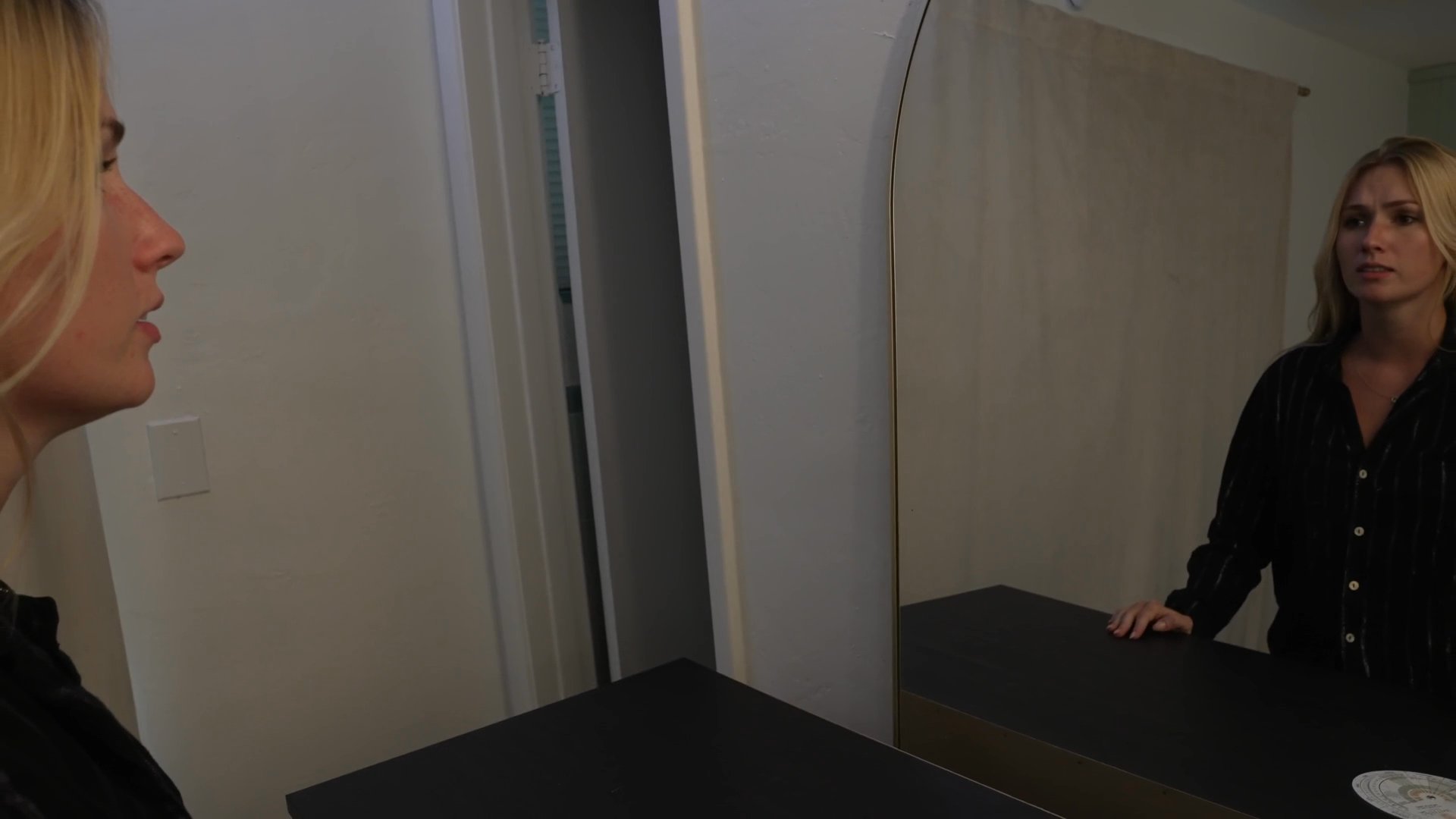 A woman wakes up in her bedroom and walks to a mirror to inspect her reflection. She gazes at herself intently, pausing to observe her own appearance in the dim light.