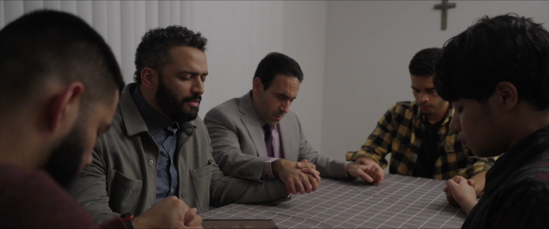 A diverse group of men sits together in a circle, holding hands and bowing their heads in collective prayer. The scene transitions to a focus on one man as he leads the contemplative moment.