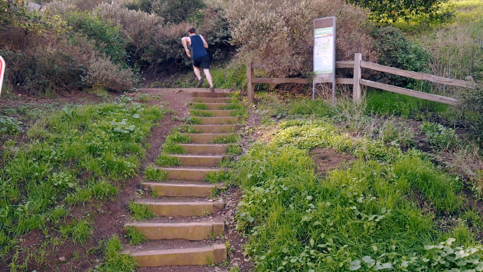 A man engages in cardiovascular training by running through a residential street and ascending a set of outdoor stairs in a park. He maintains a consistent pace while navigating the hilly, grassy terrain.