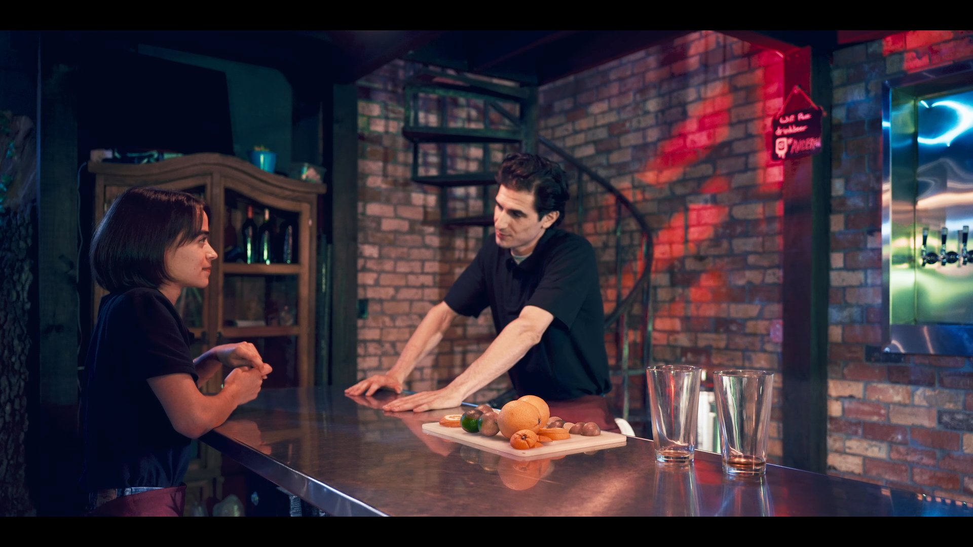 A man and a woman engage in a focused conversation at a bar counter, followed by a transition to an aerial view of a suburban neighborhood.