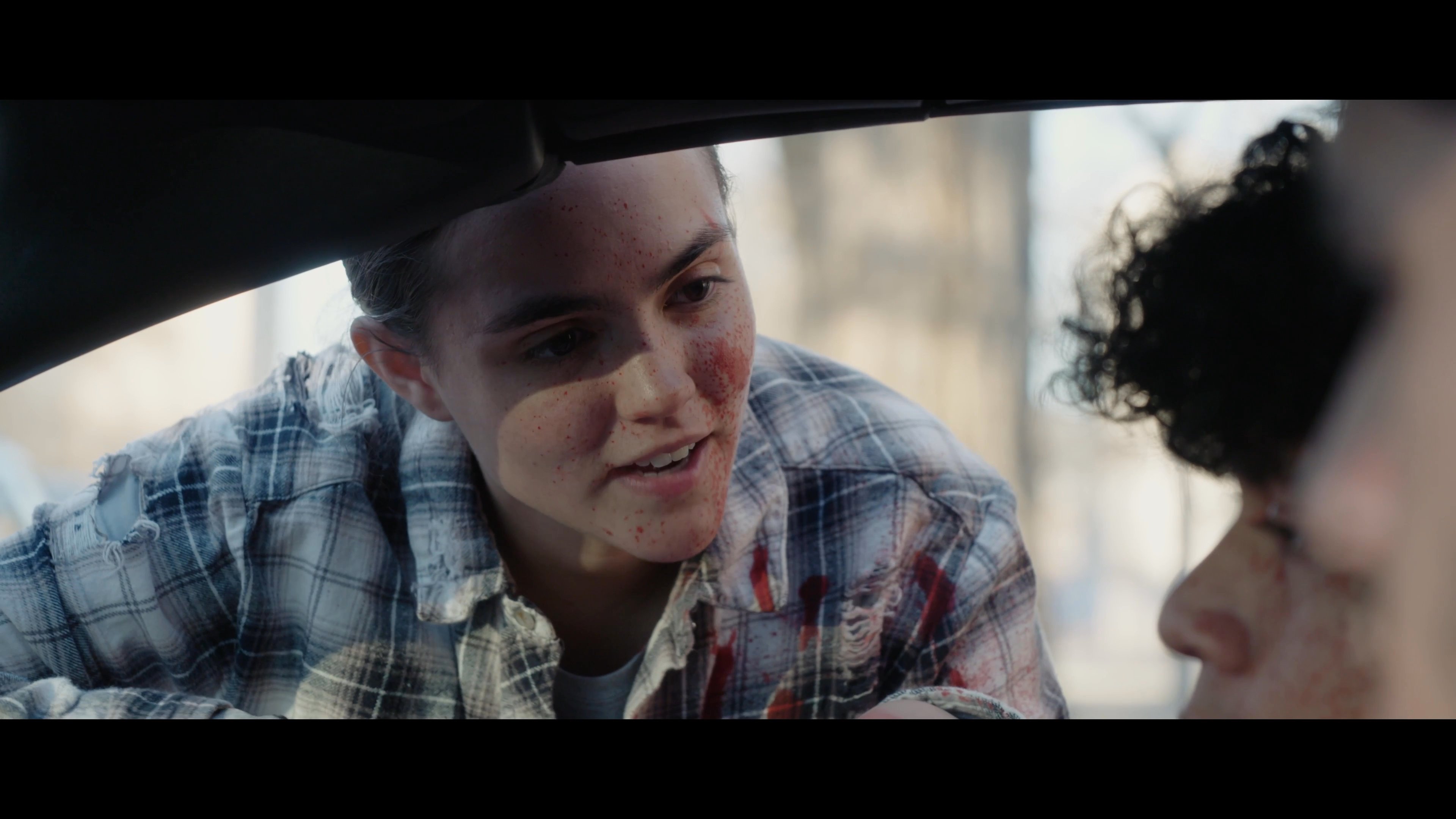 A young woman with visible injuries on her face speaks with two young men sitting inside a parked vehicle. The interaction carries a serious and intense atmosphere as they engage in a direct, focused conversation.