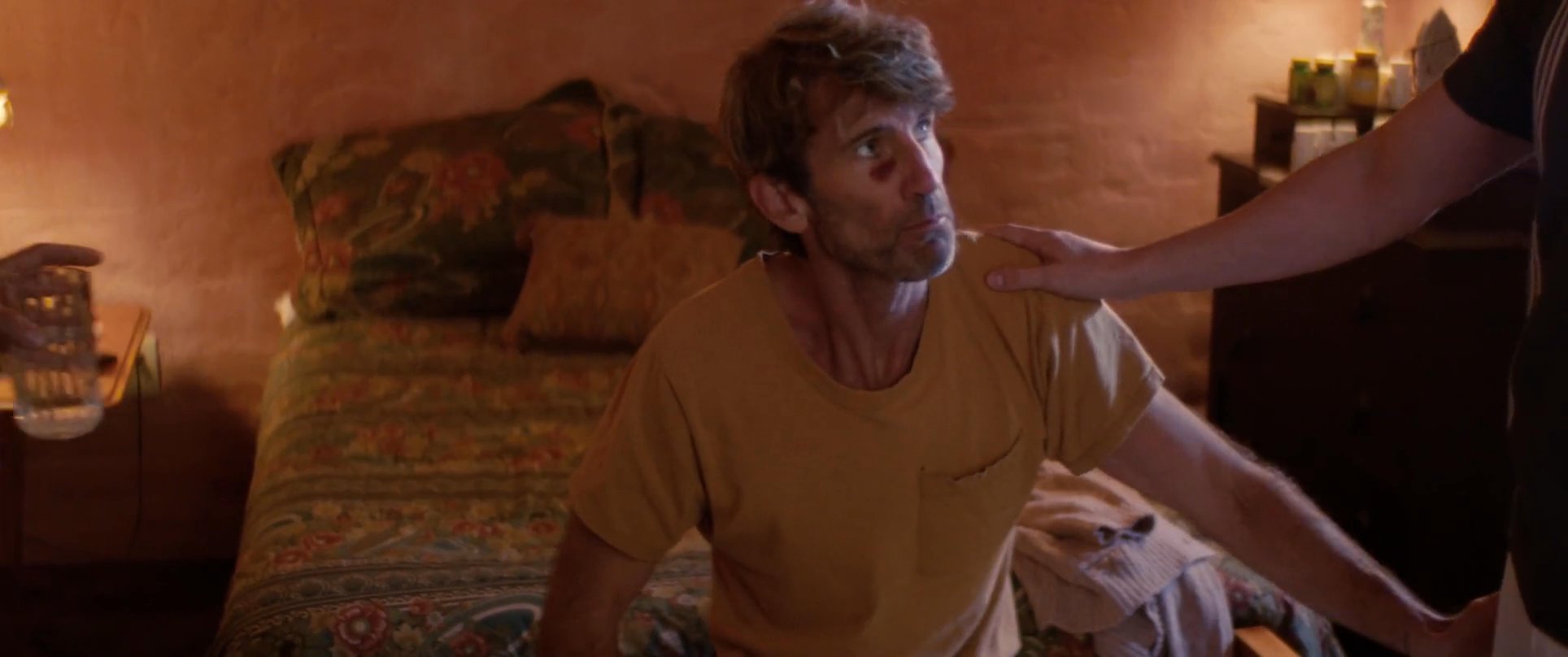 A man provides gentle physical support and assistance to another person sitting on a bed in a softly lit bedroom. The interaction highlights a moment of care, comfort, and quiet companionship between the two individuals.