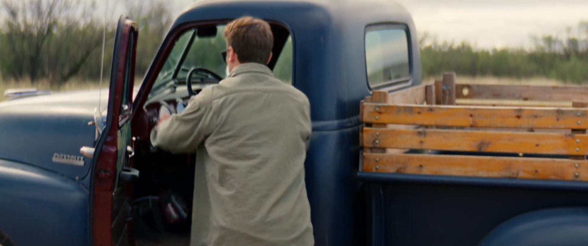 A man in a green shirt and sunglasses approaches a vintage blue pickup truck parked in a desert landscape and enters the driver's seat. The vehicle sits prominently in front of an old, weathered stone building and a windmill.