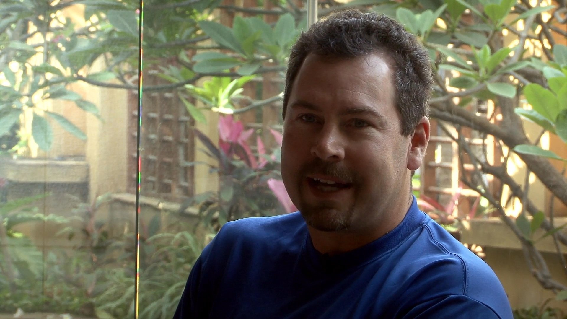 A man in a blue shirt participates in an interview while seated on a sofa in front of a garden view.